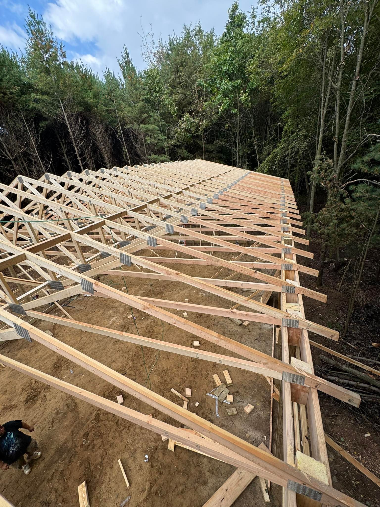 Long wooden roof trusses laid out on a dirt hillside beside dense trees and forest