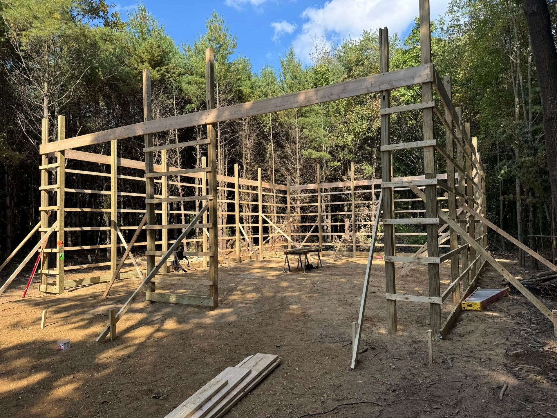 Wooden frame under construction in a wooded clearing, with ladders, braces, and scattered lumber.
