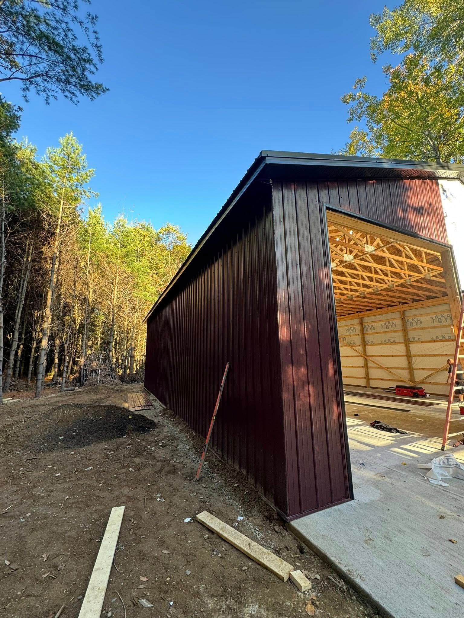 Small dark red shed under construction with open side, plywood framing, and dirt clearing surrounded by trees.