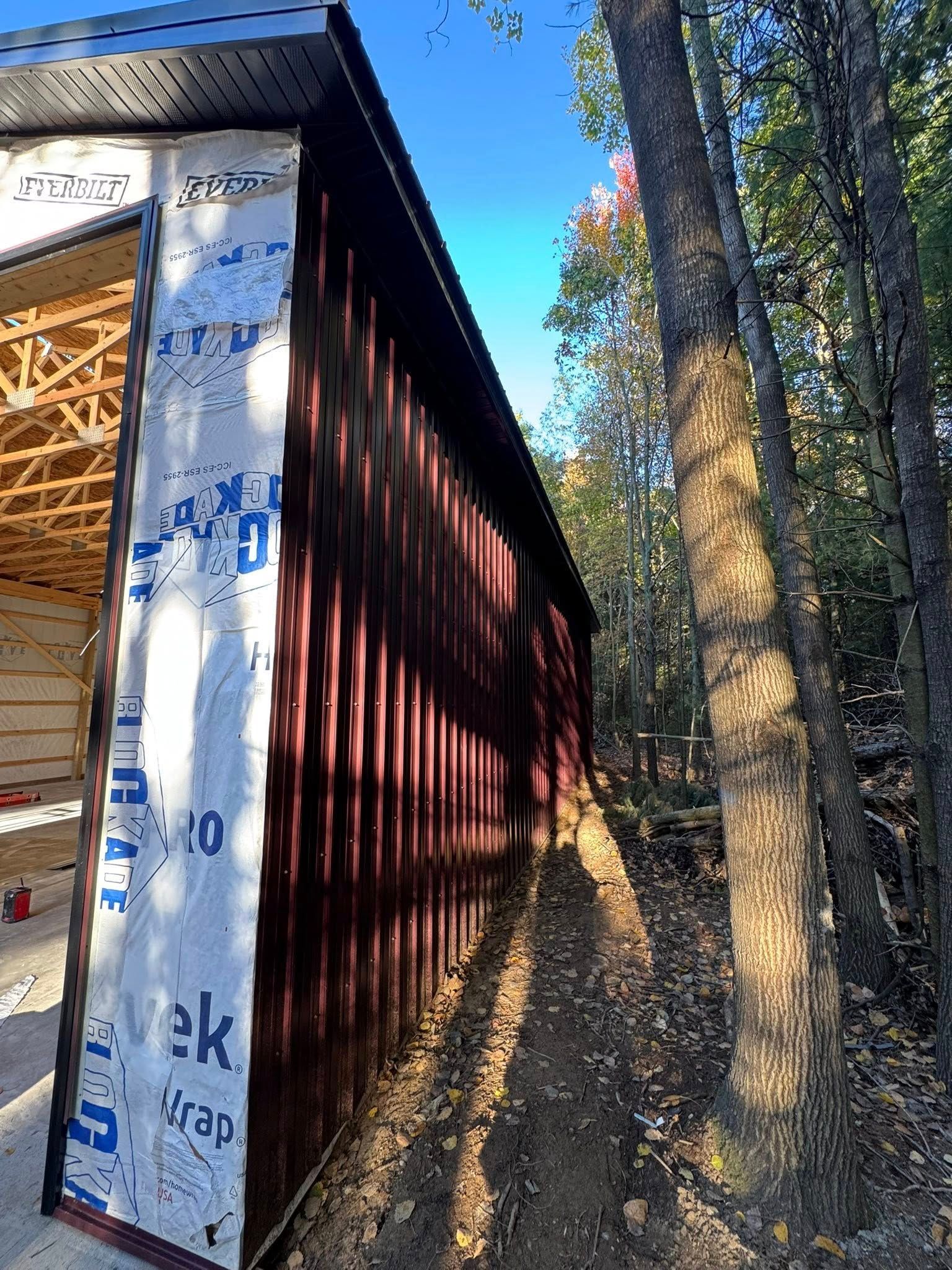 Partially built house with exposed wood framing and wrapped exterior walls beside a wooded path
