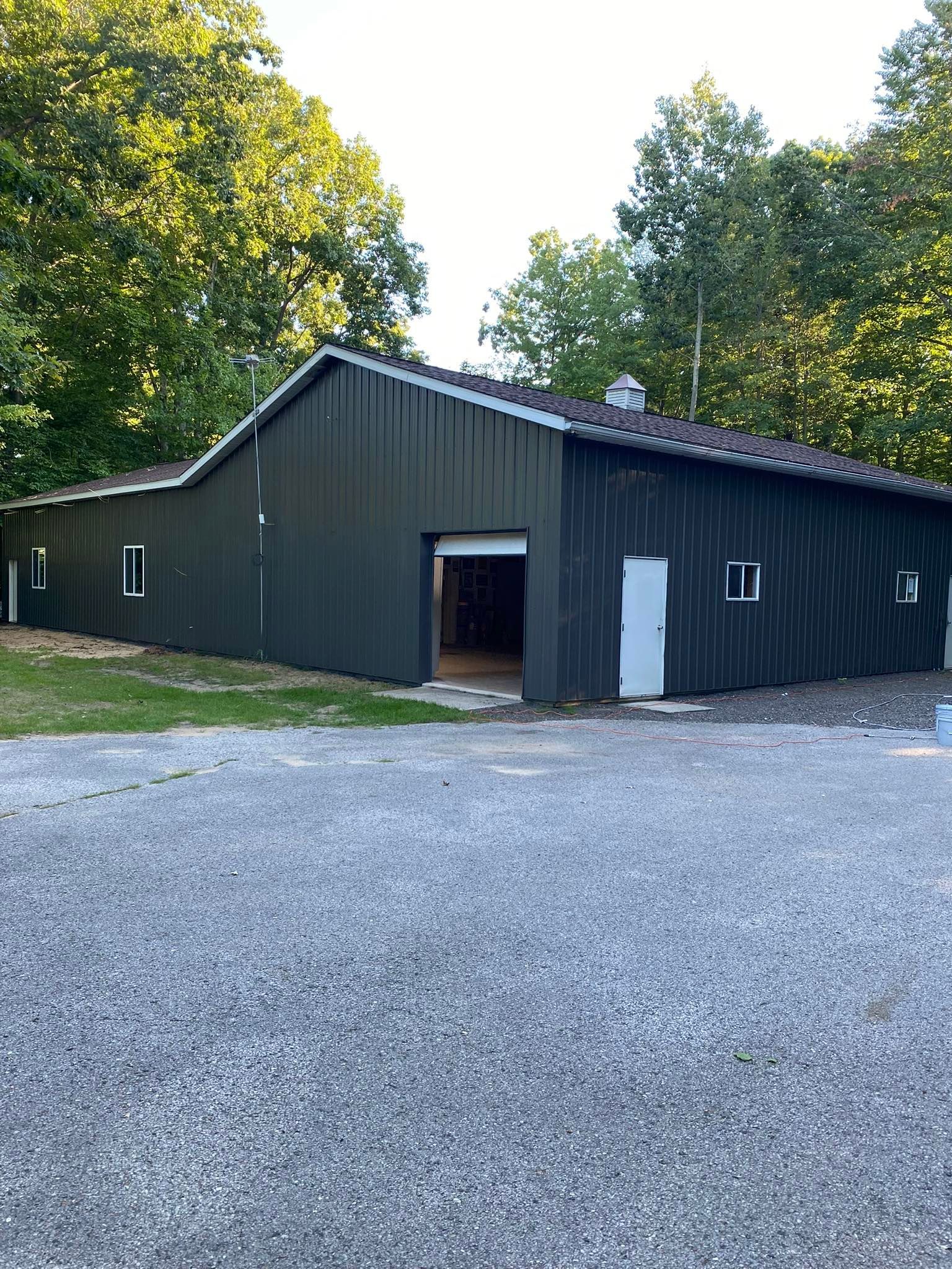 Dark green metal building beside a gravel lot and trees, with an open doorway and white side door.