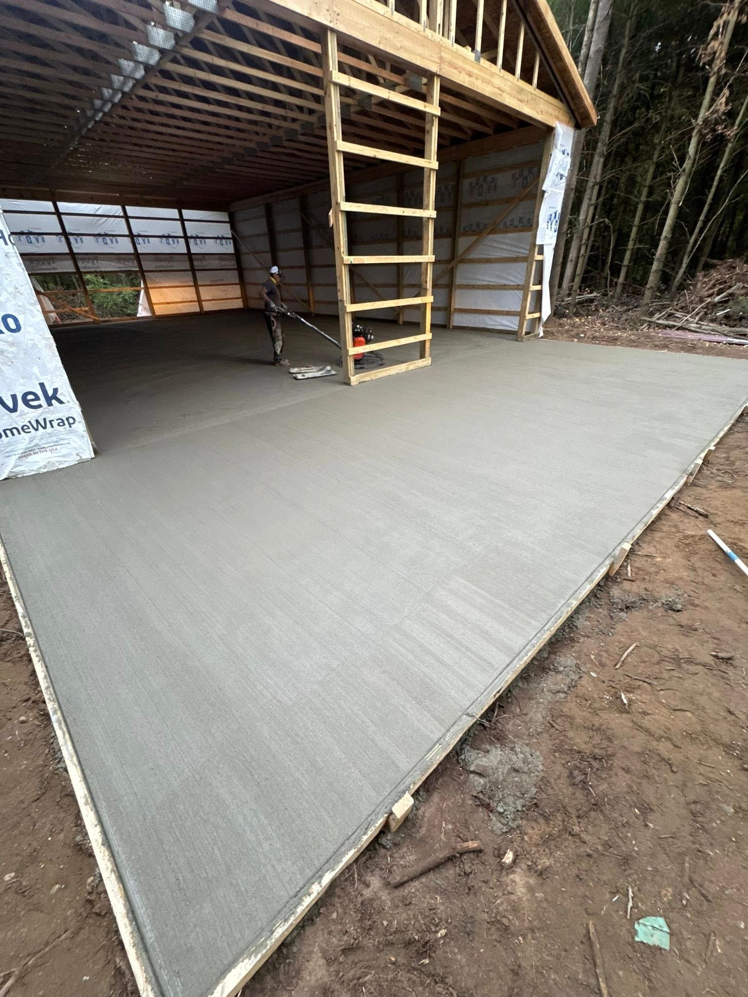Freshly poured concrete slab under a roofed structure, with a wooden ladder and formwork at one side.