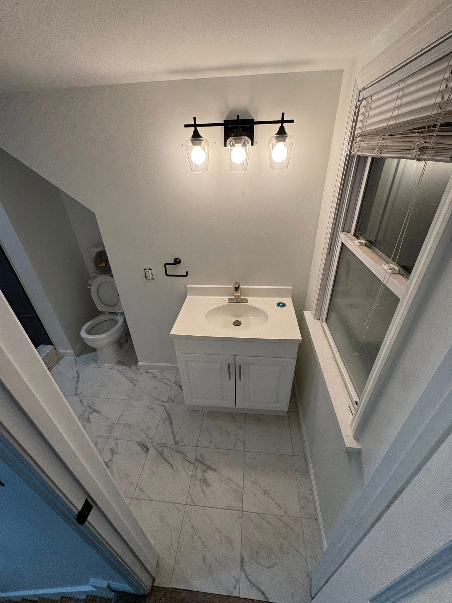Small bathroom with white vanity, toilet, marble tile floor, and window blinds.