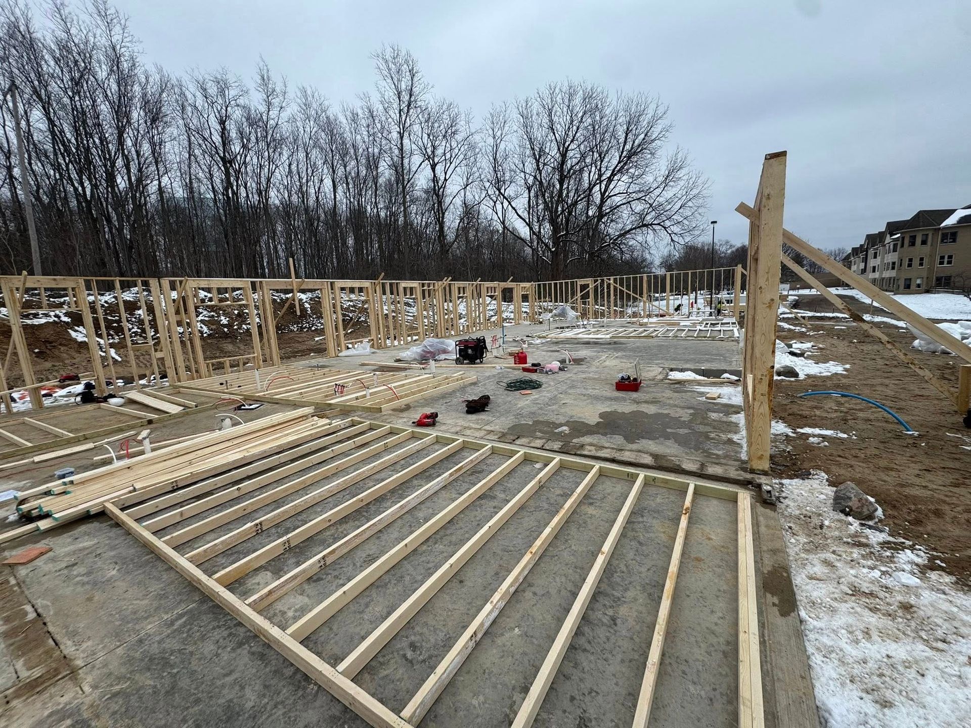 Partially built wooden frame on a snowy construction site with scattered materials and bare trees.