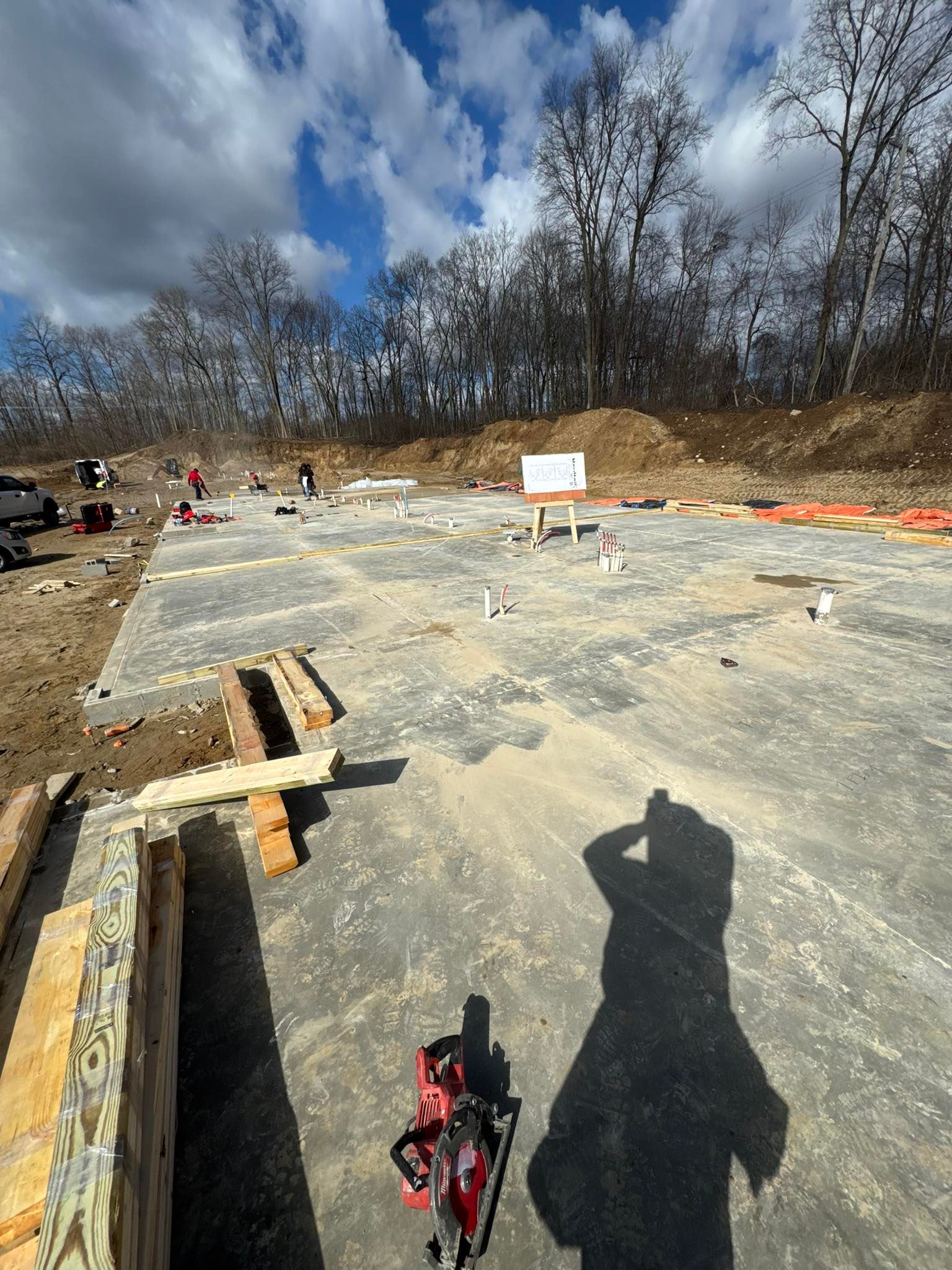 Construction site with foundation trenches, scattered materials, and cloudy sky; shadow of a person in foreground