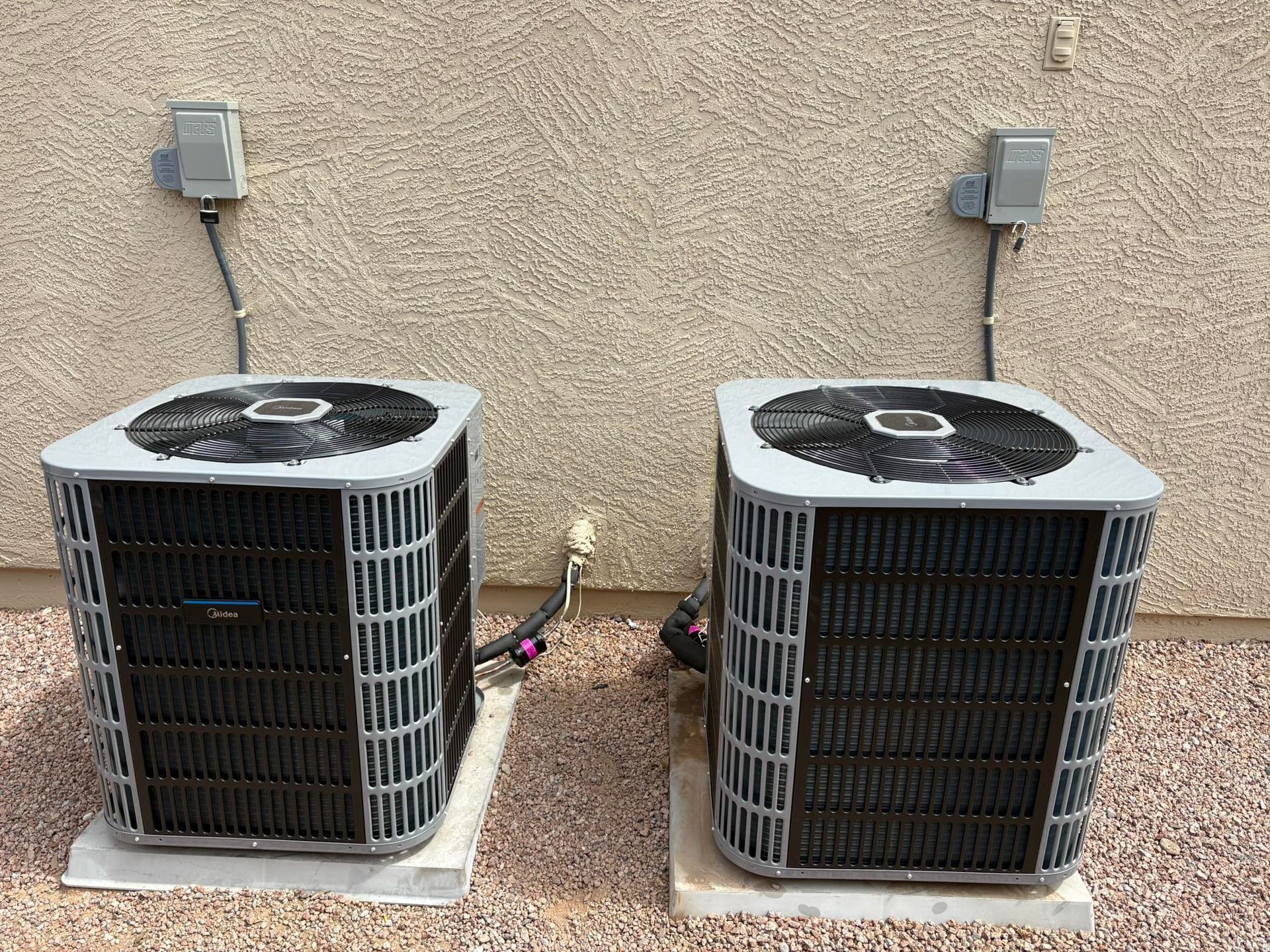 Two outdoor air conditioner units against a beige stucco wall on gravel.