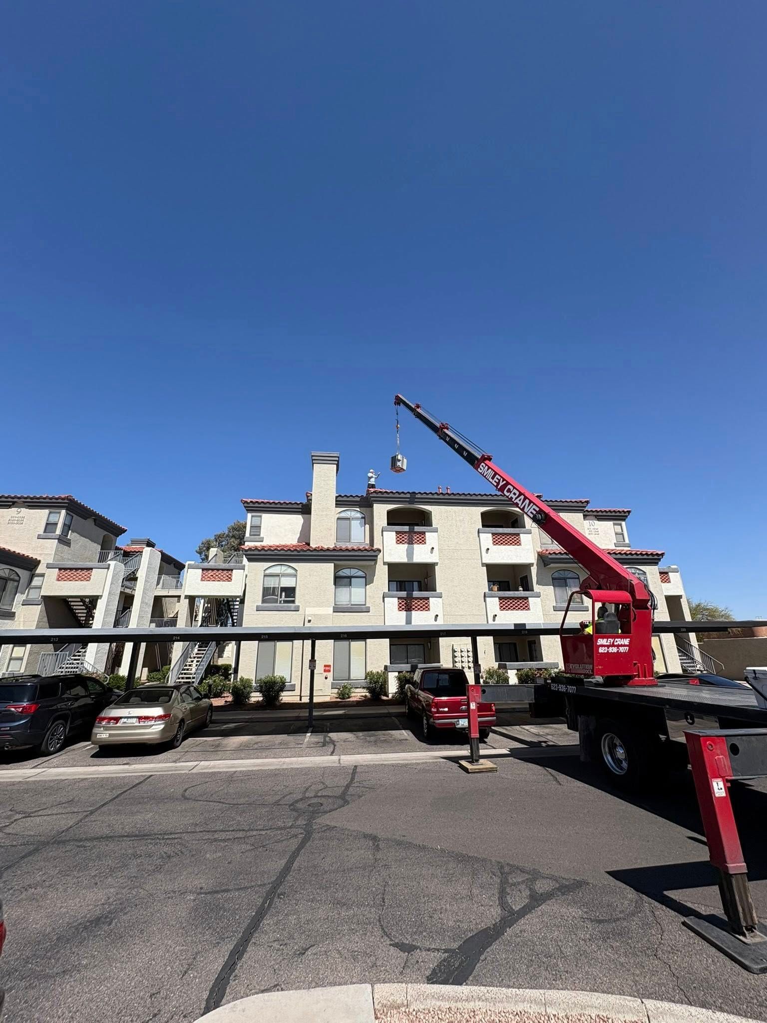 Crane lifting a red object in a parking lot beside apartment buildings under a clear blue sky