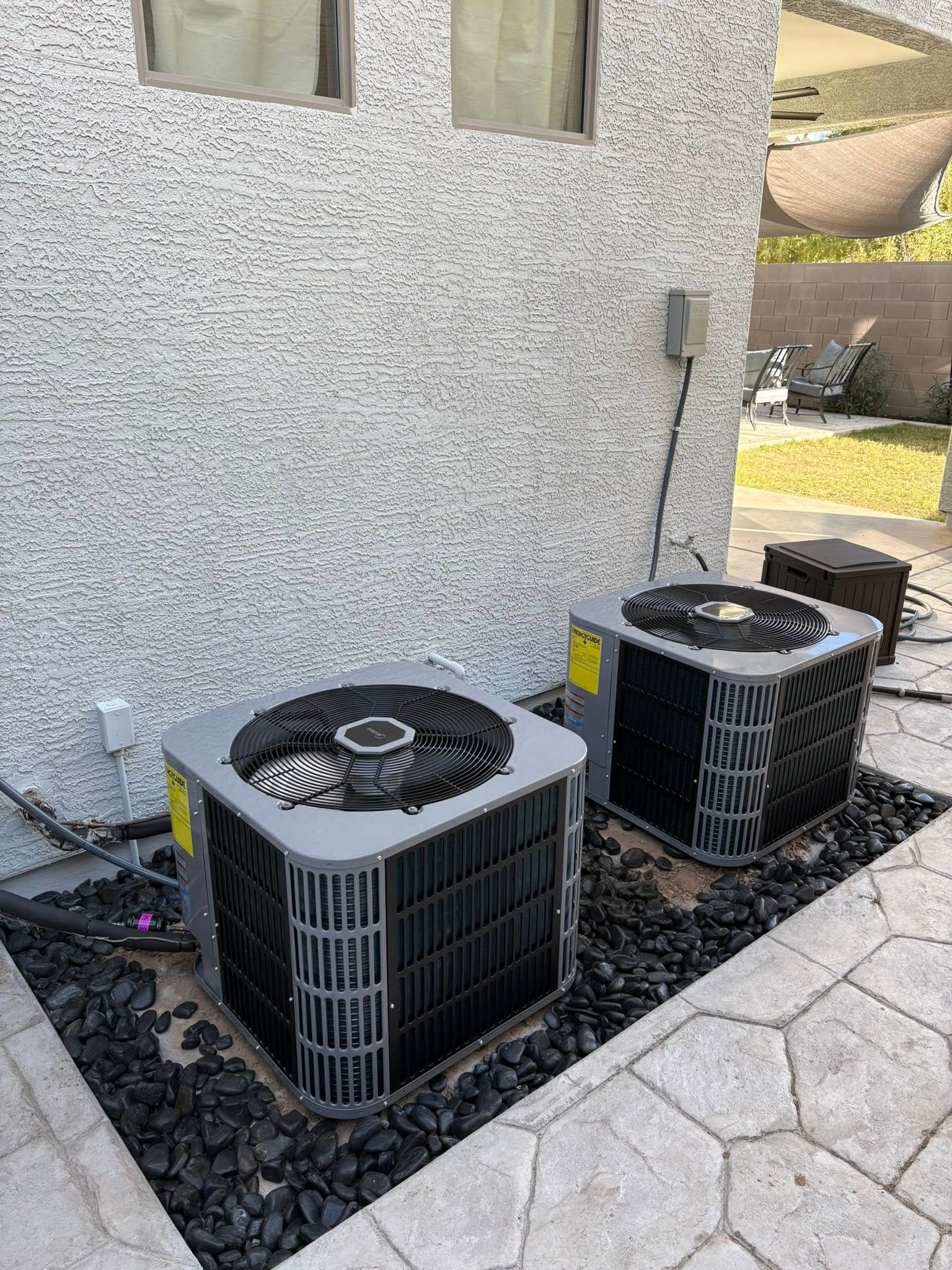 Two outdoor air conditioning units beside a stucco house wall on a gravel bed next to a paved walkway