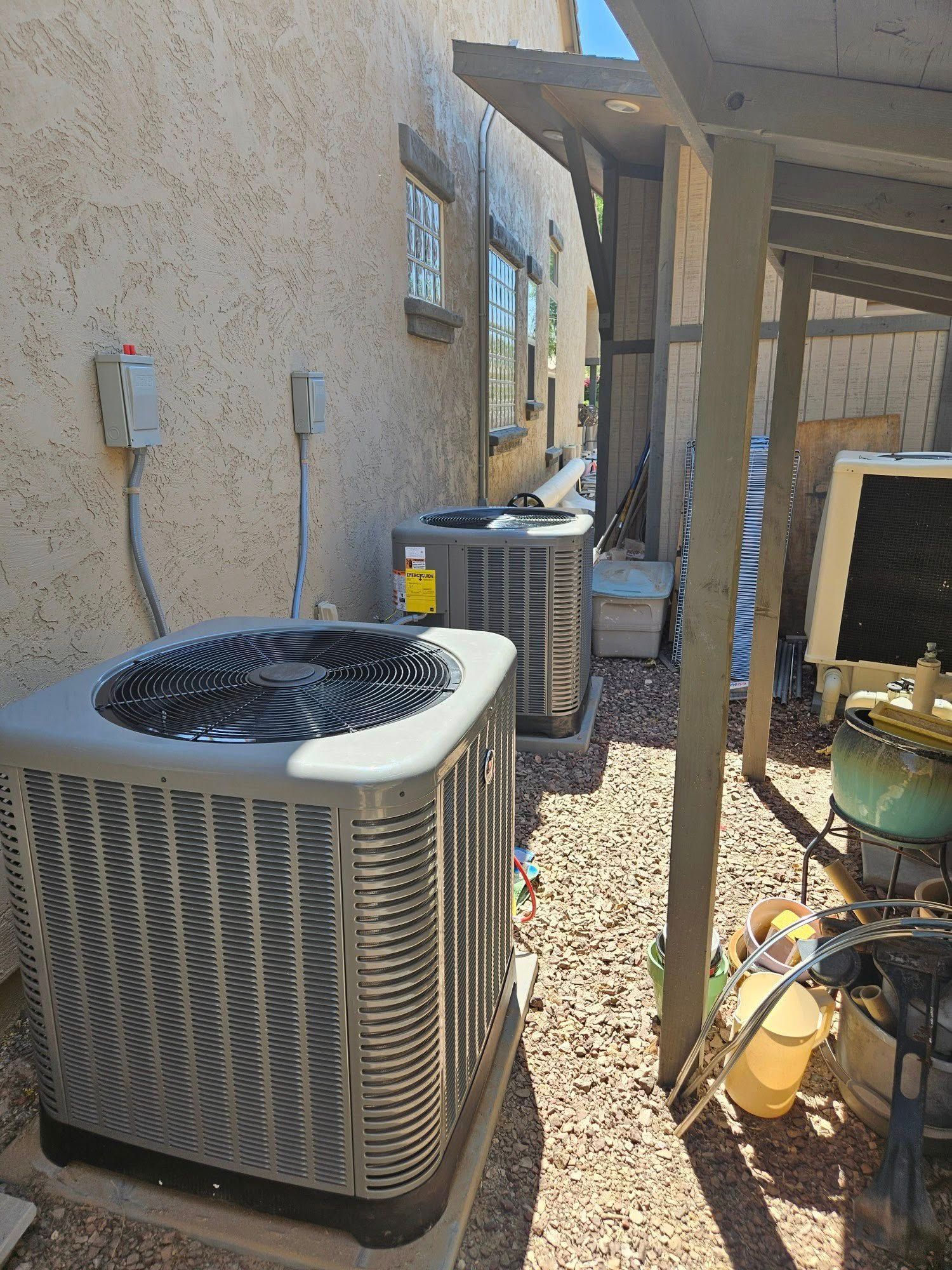 Outdoor HVAC units beside a stucco house, with a shaded patio and gravel ground.