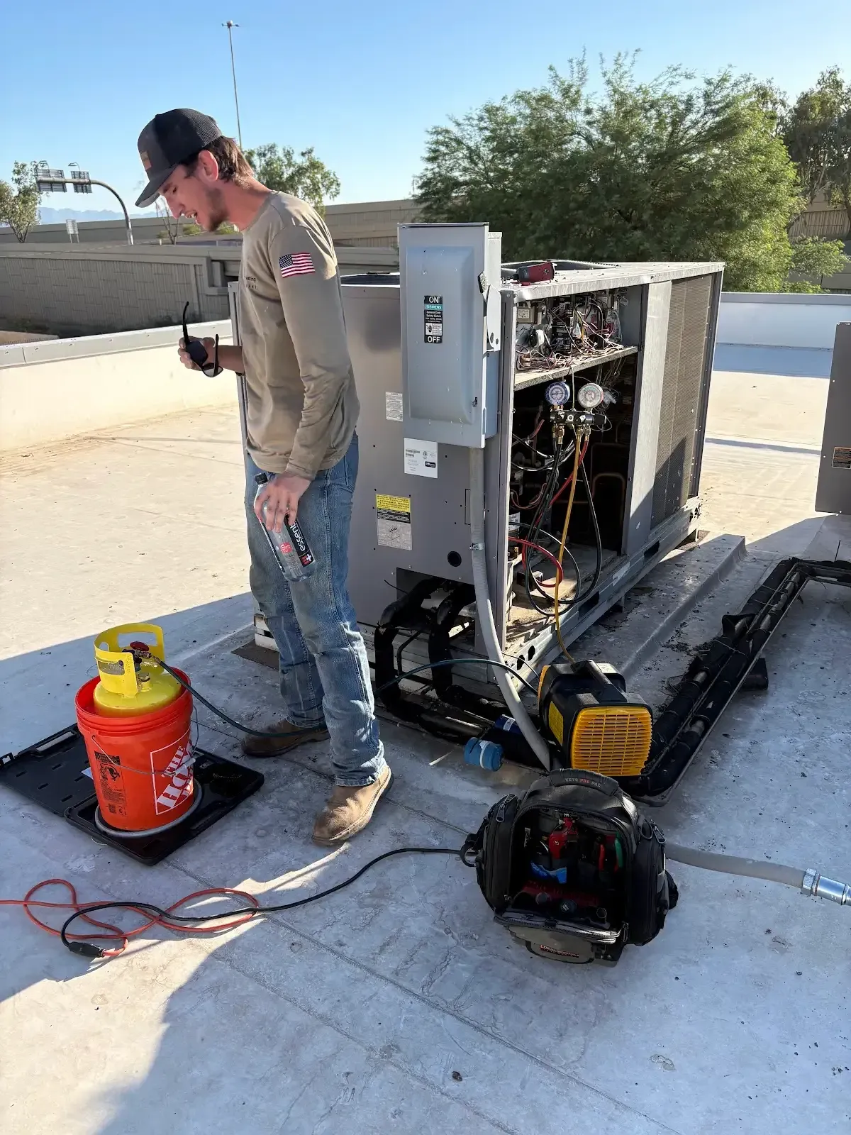 Worker beside exposed HVAC unit on a rooftop, with tools and a red refrigerant tank nearby.
