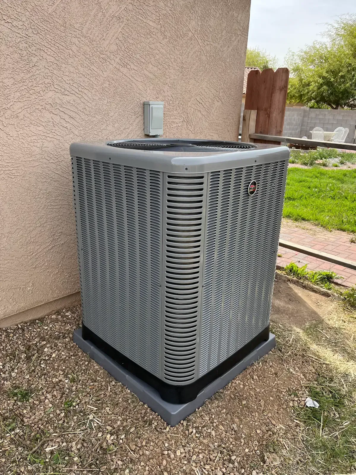 Gray outdoor air conditioning unit beside a house wall on a gravel patio, with grass and fence in background