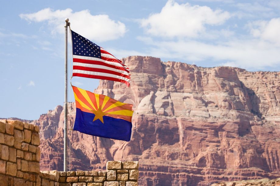American and Arizona flags fly in front of a red rock cliff under a blue sky.