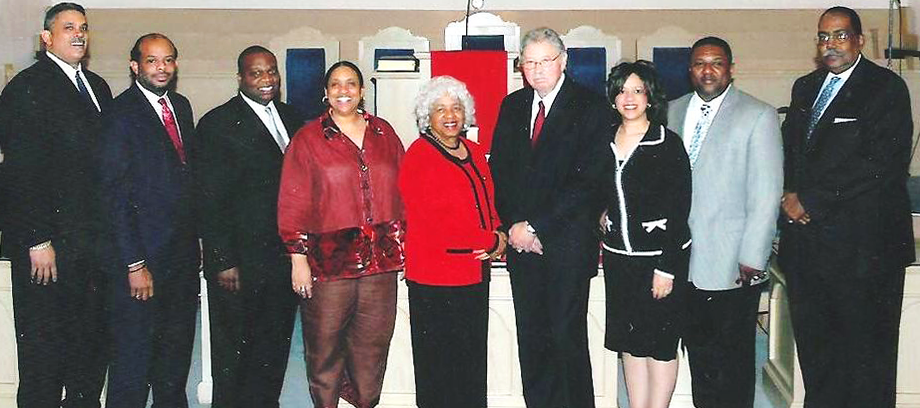 Nine people in formal attire standing together indoors at a ceremony, with one woman in a red jacket centered.