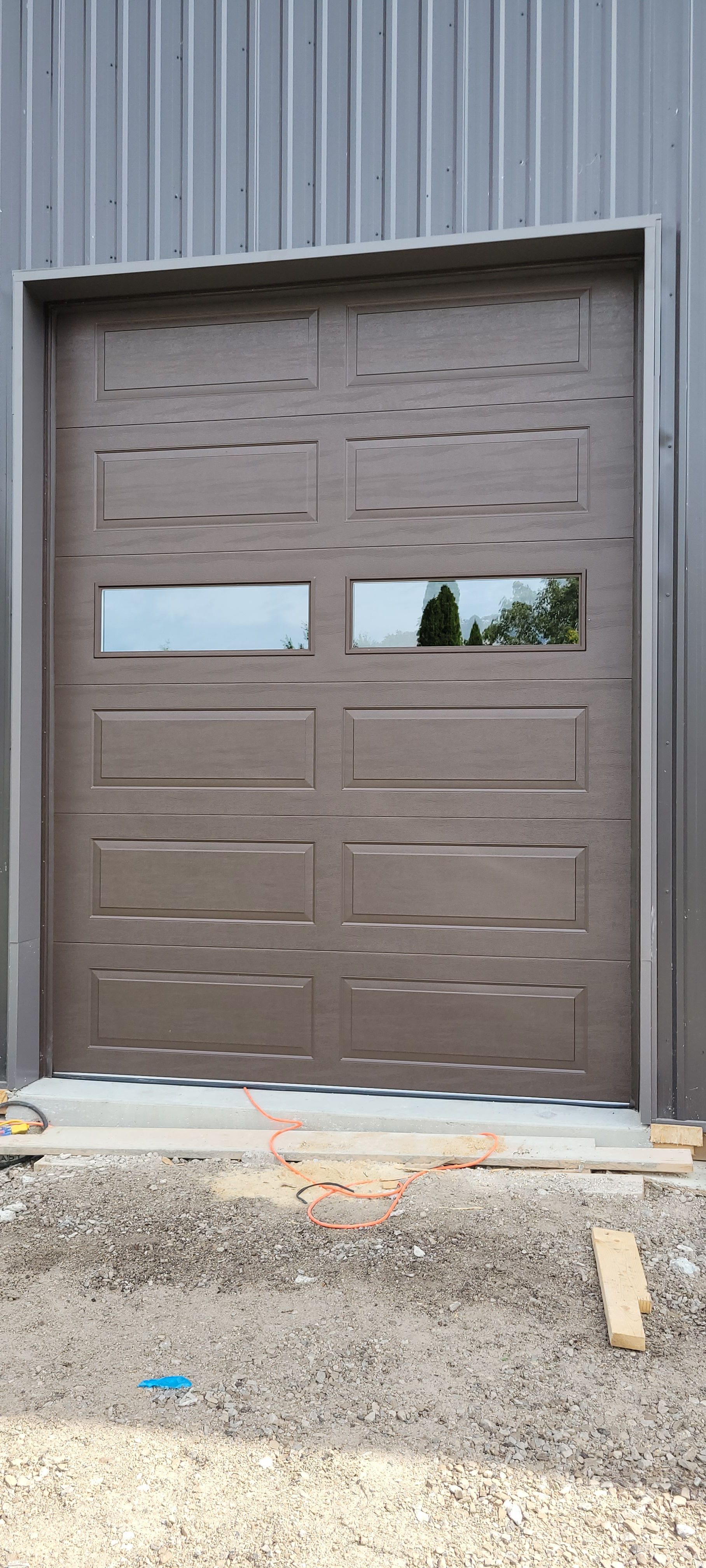 A brown garage door with two rectangular windows is set in a gray metal building. Crushed rock lies in front of the door.