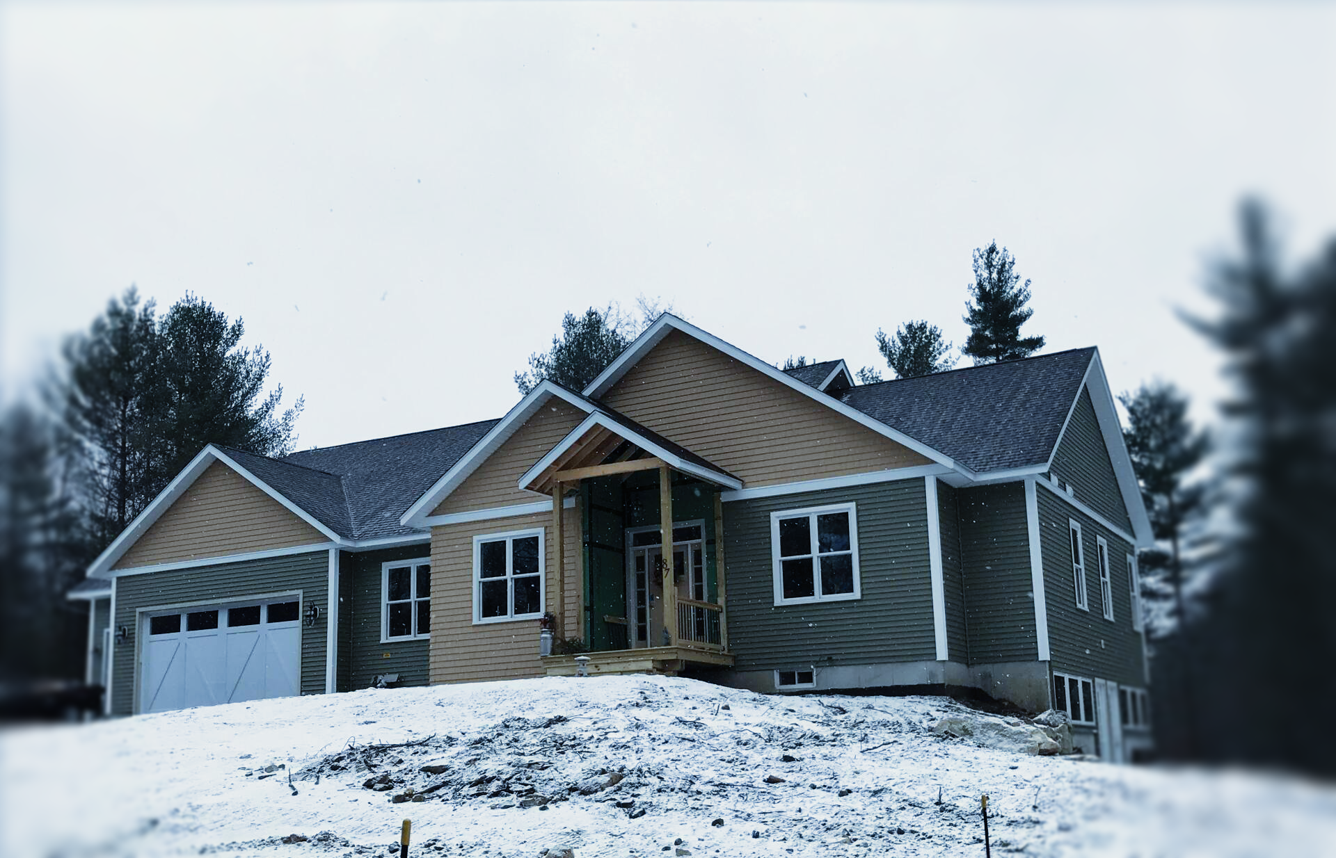 A house under construction with light wood siding and dark roofing, situated on a snow-covered hill among trees.