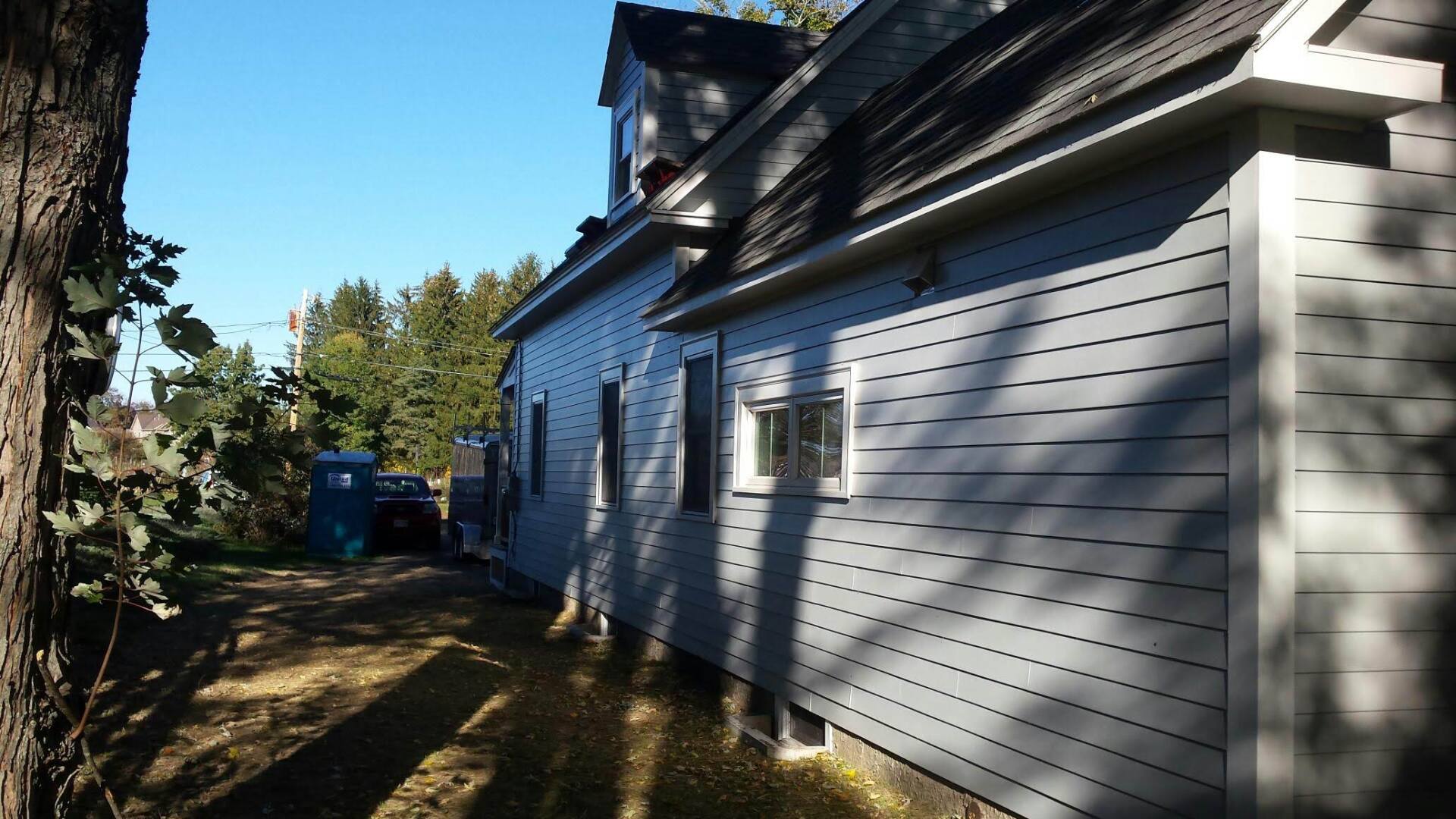 A side view of a light blue house exterior with horizontal siding, dark window frames, and trees in the background.