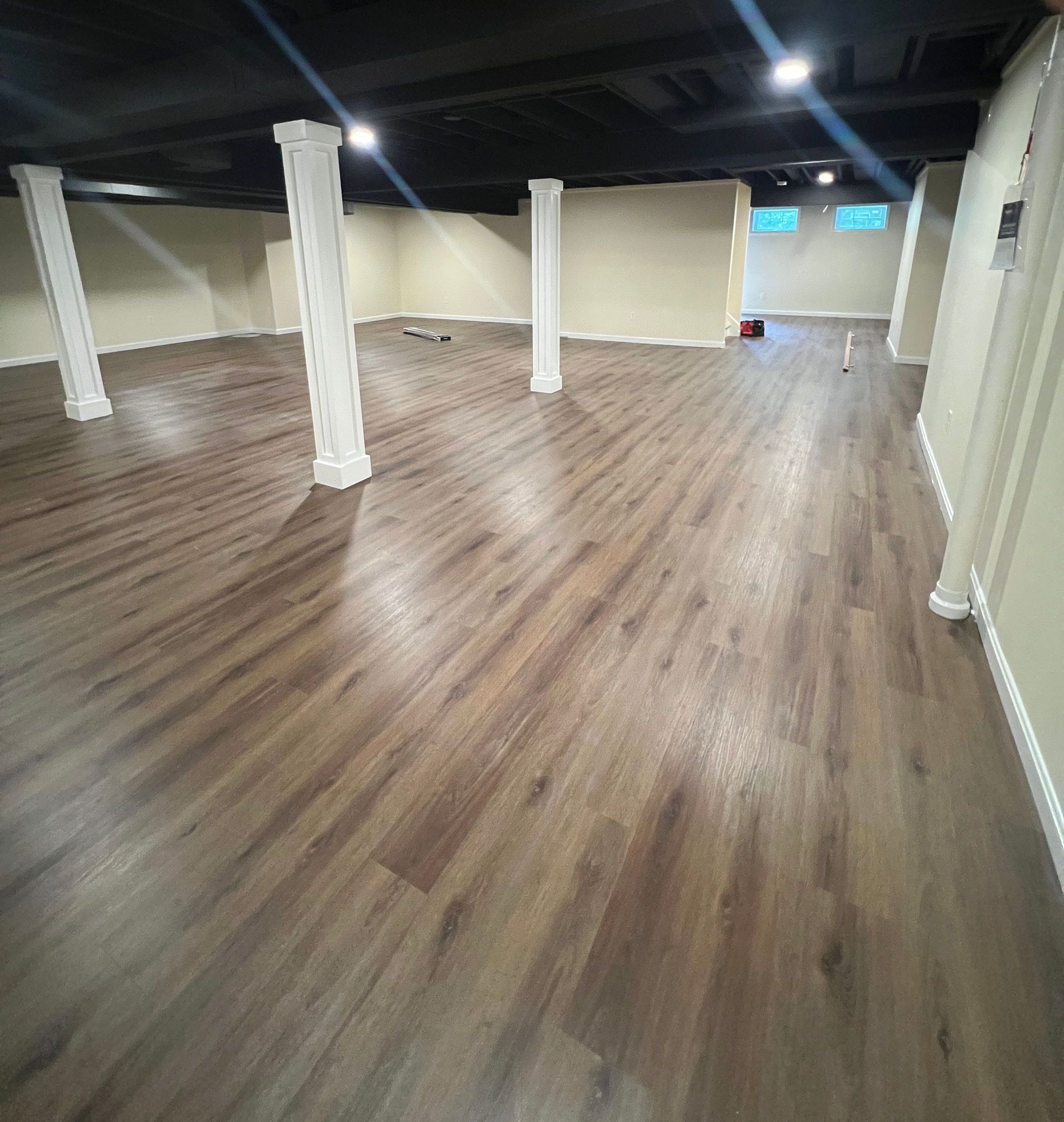 An open, unfinished basement with brown wood-look vinyl flooring, white support columns, and a painted black ceiling.