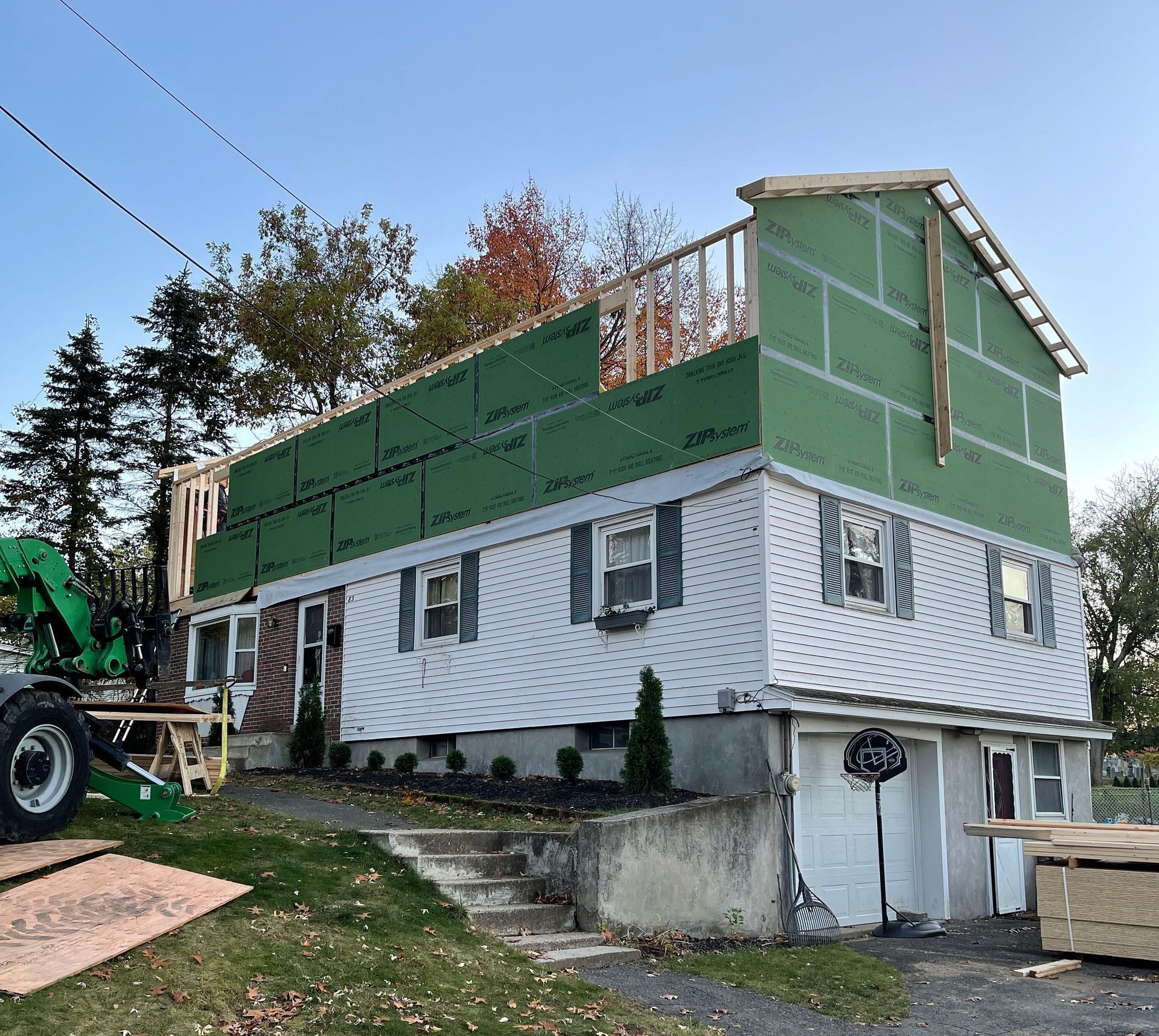A two-story house under construction with a new upper level framed and covered in green sheathing, next to a large crane.