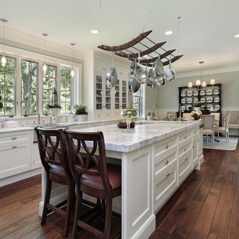 A bright, white kitchen with a large marble island, two wooden chairs, and a hanging pot rack above a wood floor.
