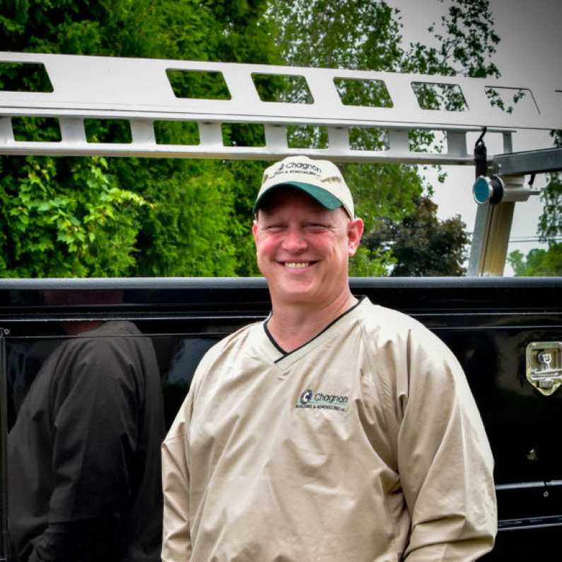 A smiling person in a tan windbreaker and green cap stands in front of a truck with a roof rack.
