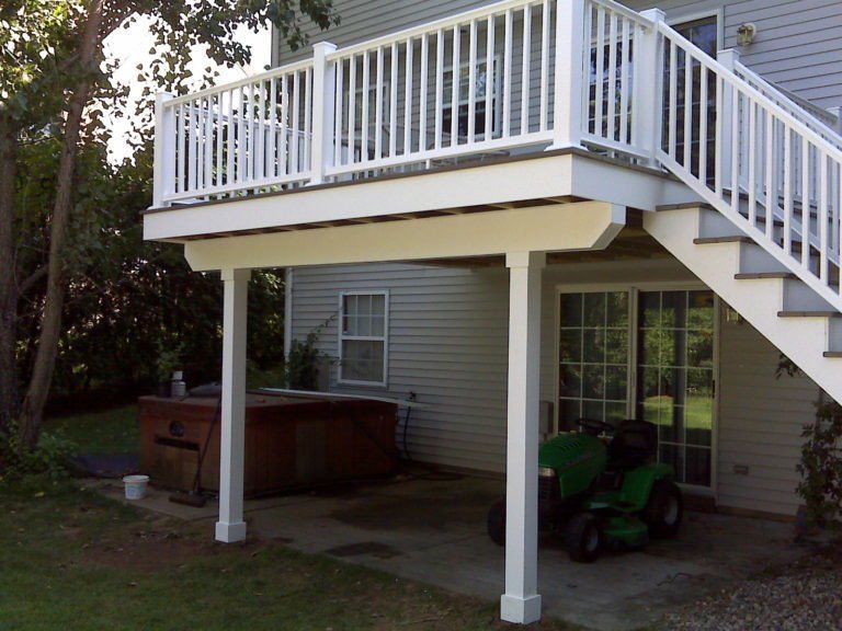 A wooden deck with white railings over a concrete patio featuring a hot tub and a green lawn tractor.
