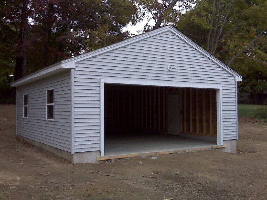 A grey, vinyl-sided detached garage with an open doorway, white trim, and concrete foundation in a wooded setting.