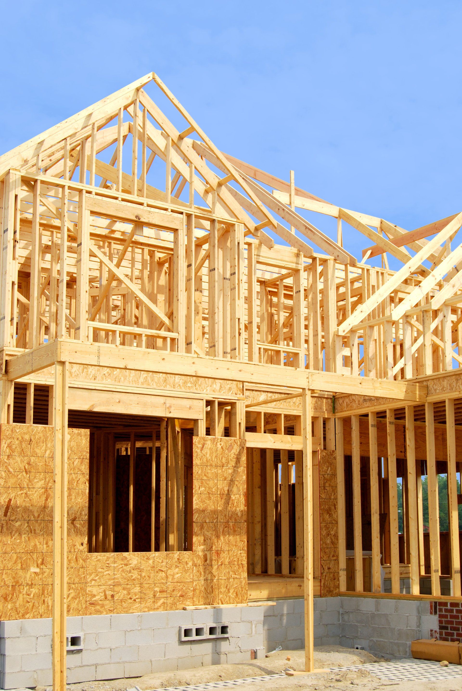 A wood-framed house under construction with visible wall studs, roof trusses, and a concrete foundation block base.