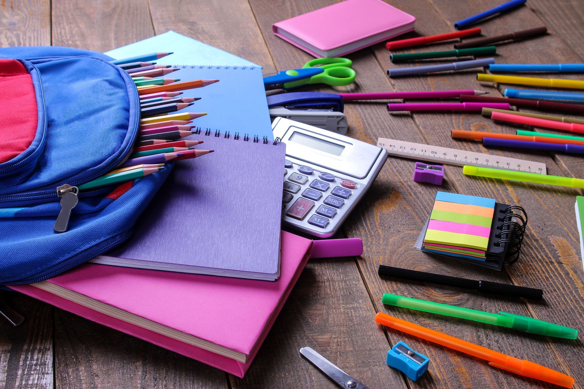 Uno zaino blu pieno di matite colorate è appoggiato su una scrivania di legno accanto a quaderni, una calcolatrice e materiale scolastico.