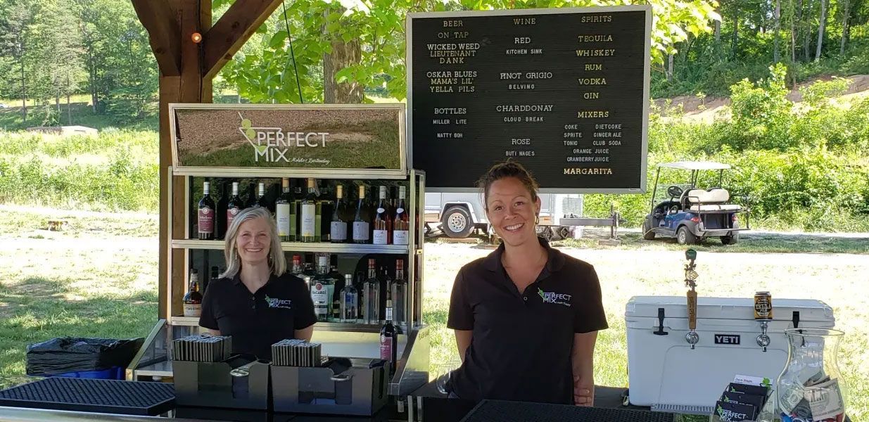 Two women are standing behind a bar in a field.