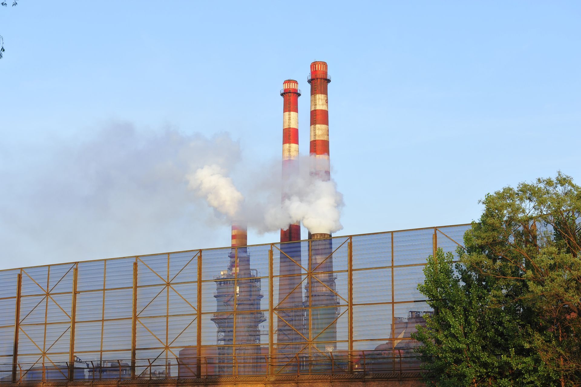 Red and white industrial smokestacks emit white smoke behind a metal fence with trees to the right under a blue sky.