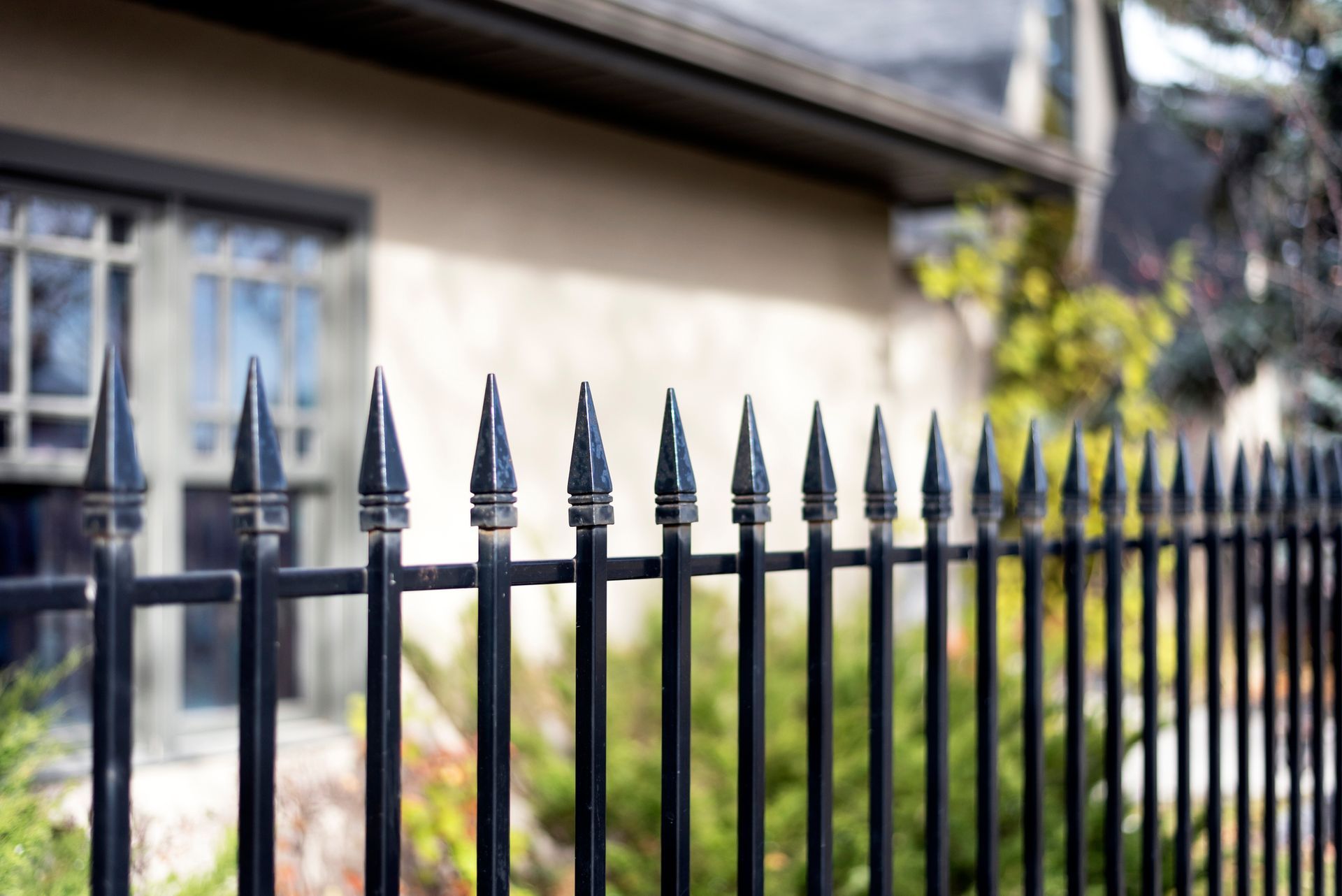 A black metal fence with pointed finials in the foreground, with a blurred house and greenery in the background.