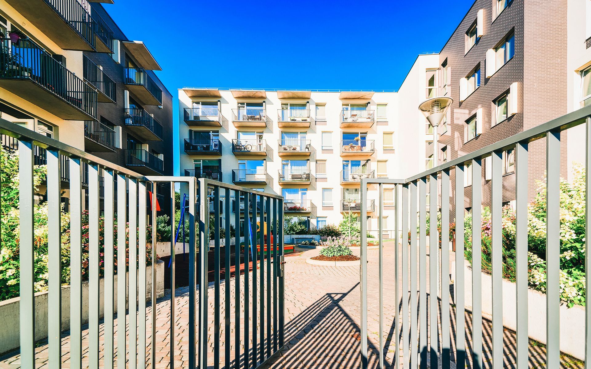 A gated entrance opens into a sunlit courtyard between three modern multi-story apartment buildings.