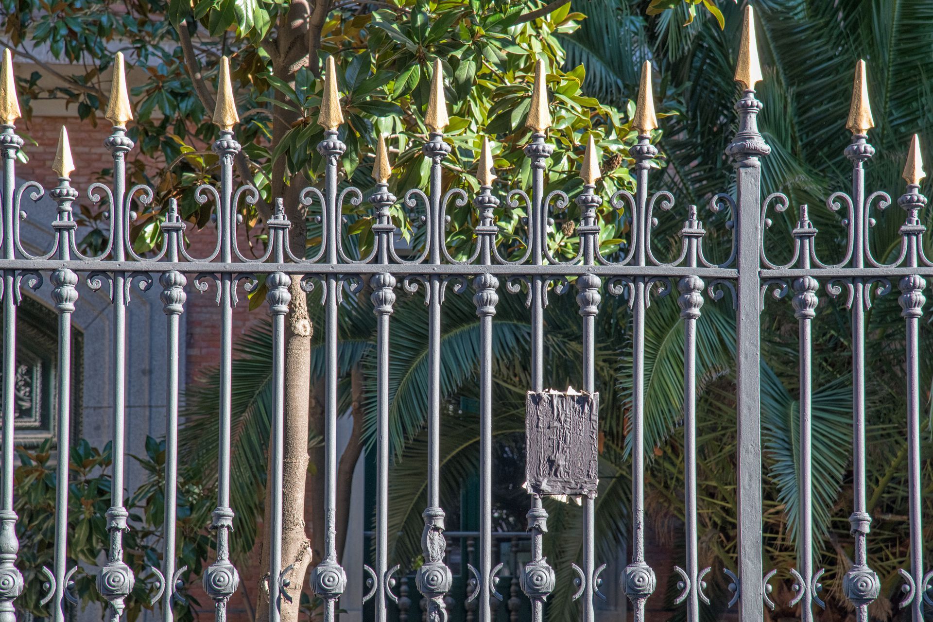 A black metal fence with gold-tipped spikes stands in front of lush green trees and palm leaves.