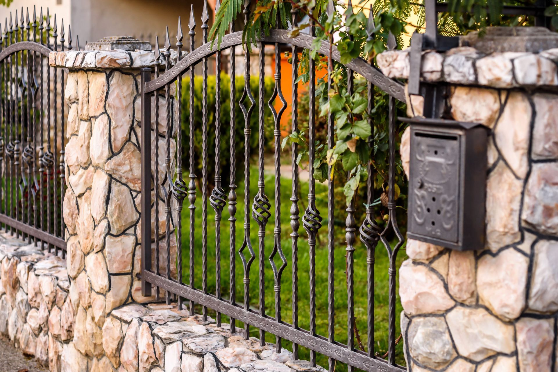 Black wrought iron fence with decorative details mounted between stone pillars, with a mailbox attached to one pillar.
