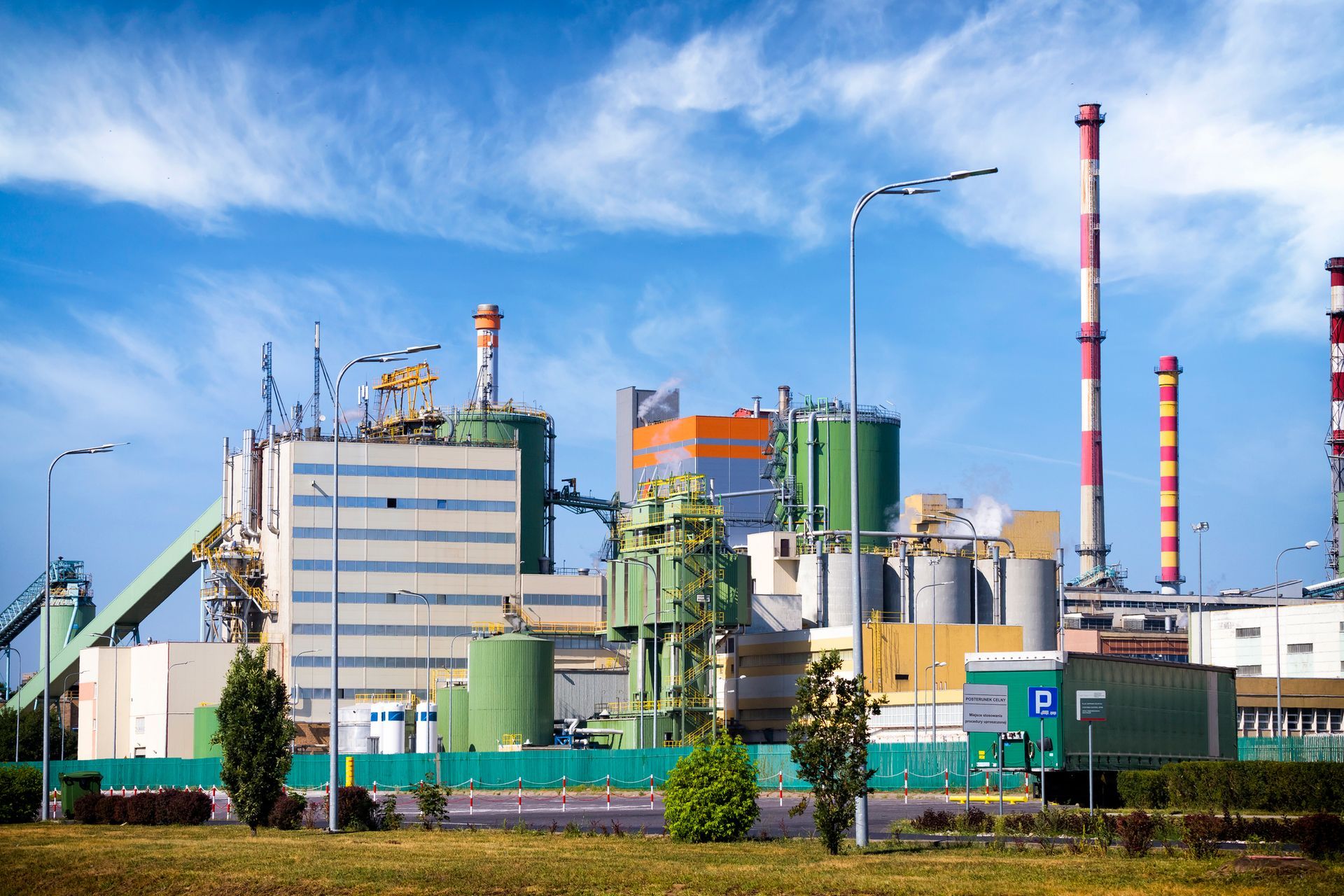 Industrial plant with green and white structures, towering red and white smokestacks, and a blue sky in the background.