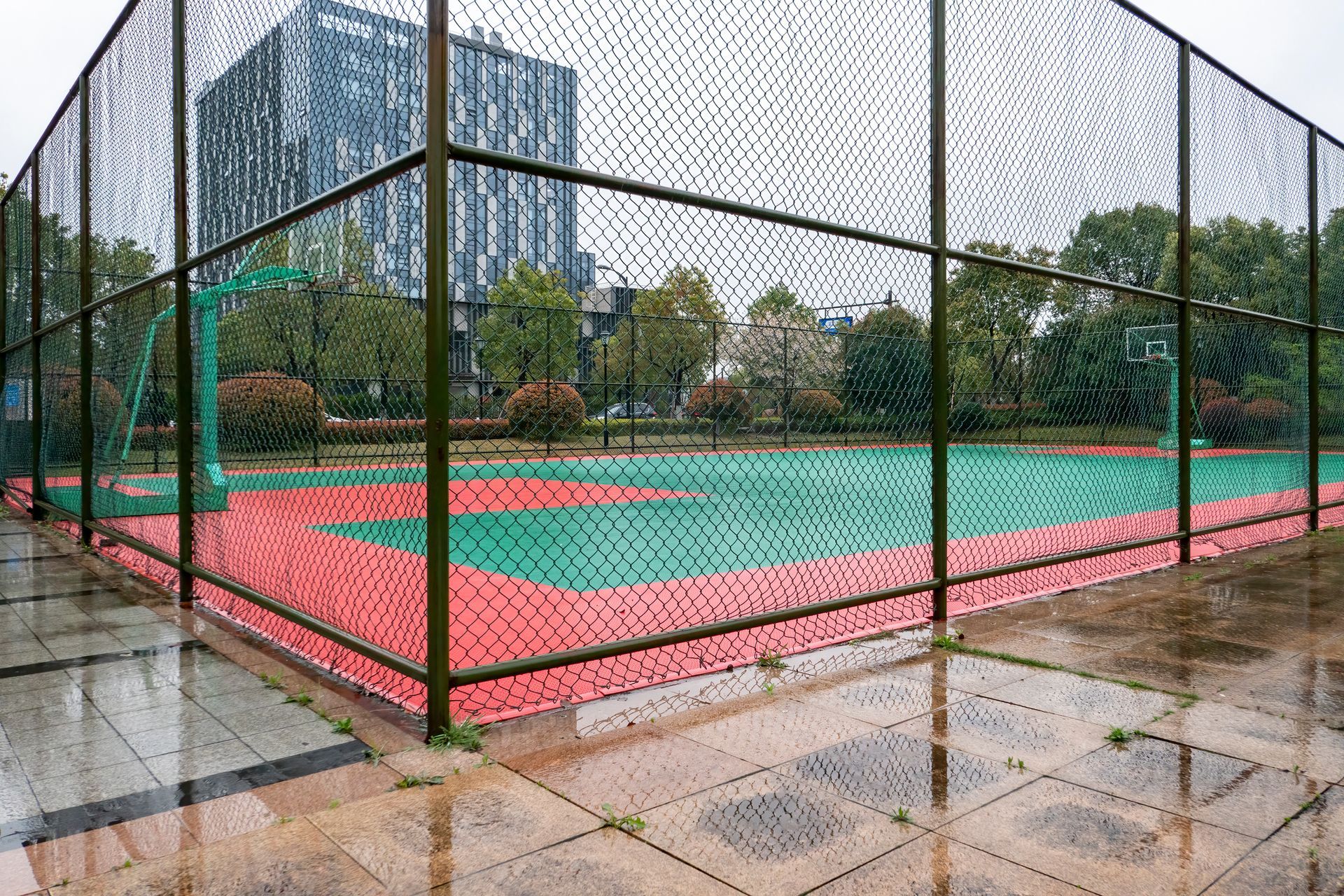 An empty outdoor basketball court enclosed by a chain-link fence on a rainy day, featuring red and green court flooring.