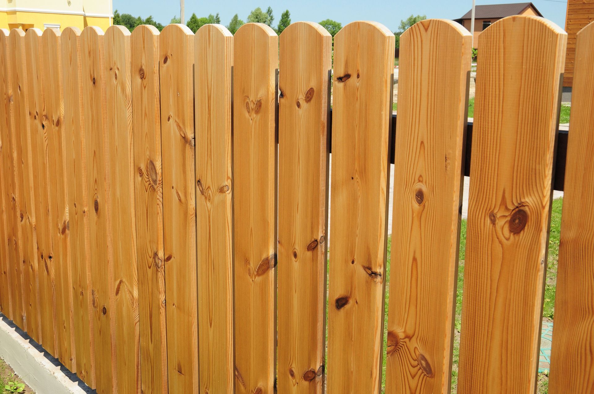 A close-up view of a wooden picket fence with a natural, light-brown finish, positioned outdoors in a residential area.