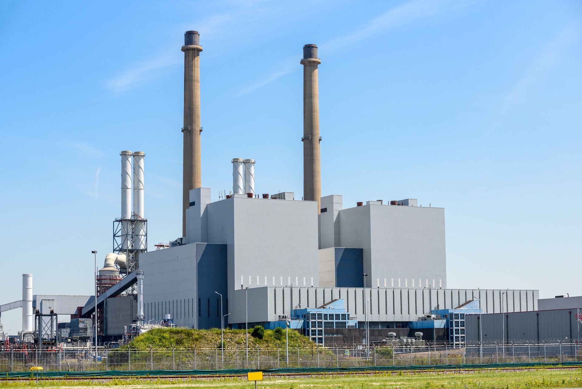 A large, modern power plant with two tall chimneys against a clear blue sky, surrounded by industrial structures.