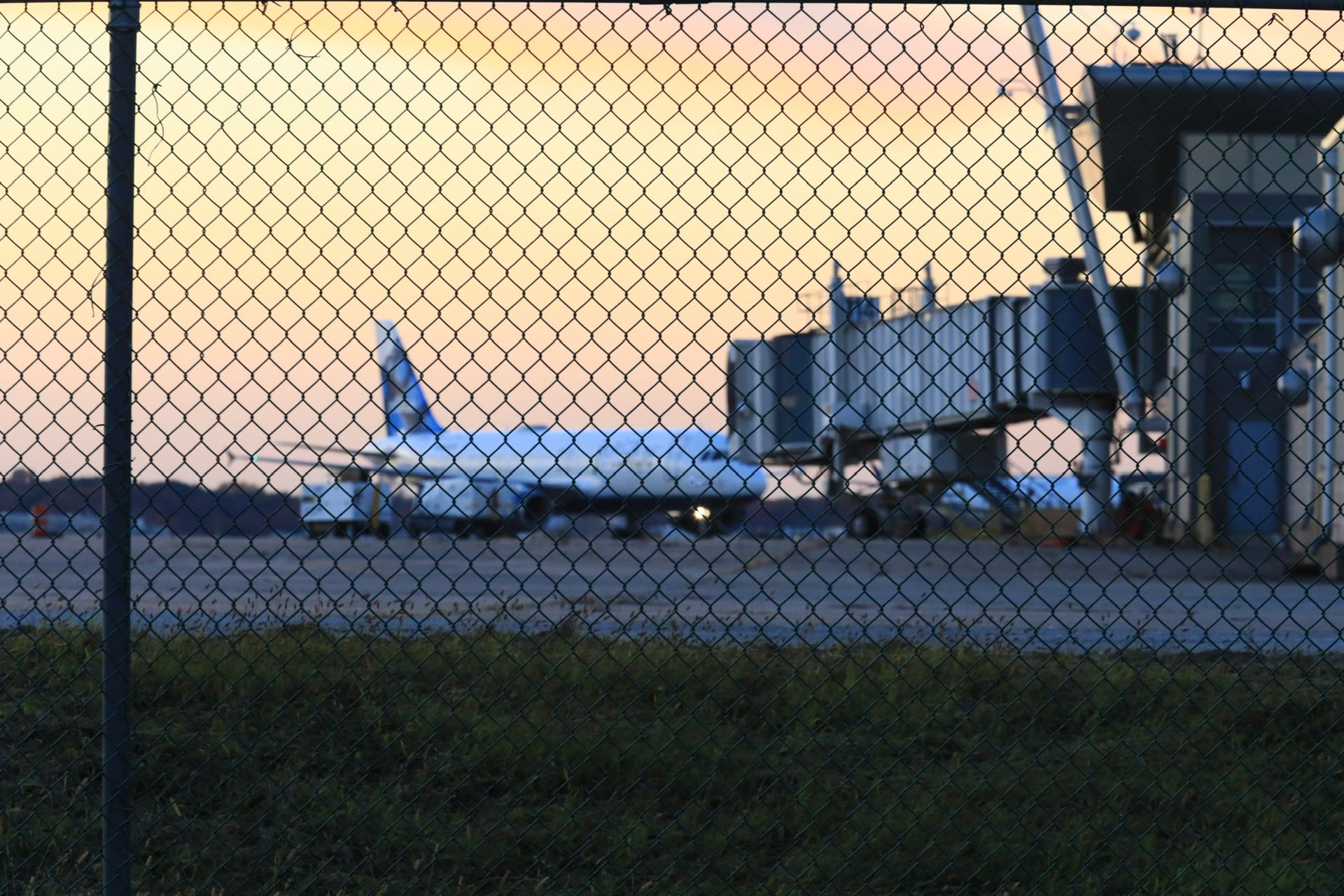 An airplane parked at an airport terminal gate, viewed through a chain-link fence at sunset.