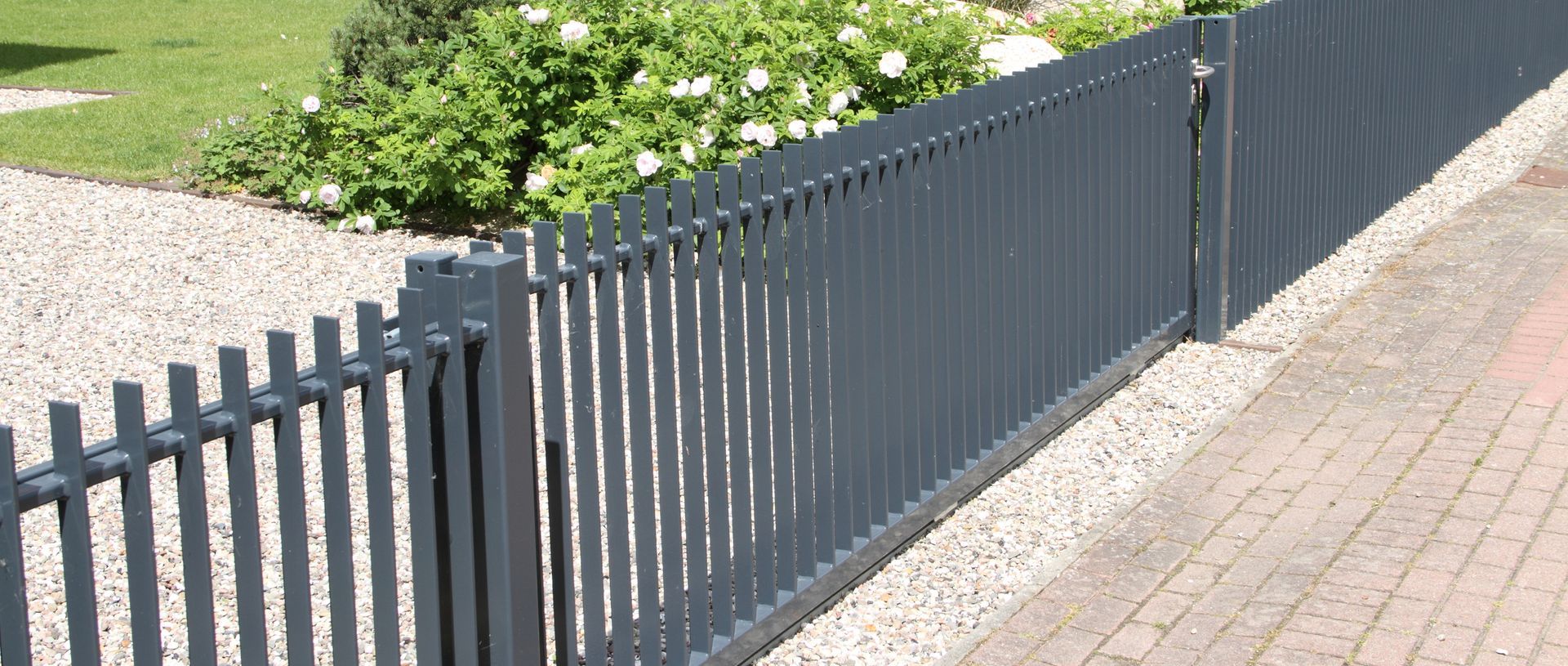 A grey metal picket fence separates a gravel driveway and green shrubbery from a brick sidewalk.