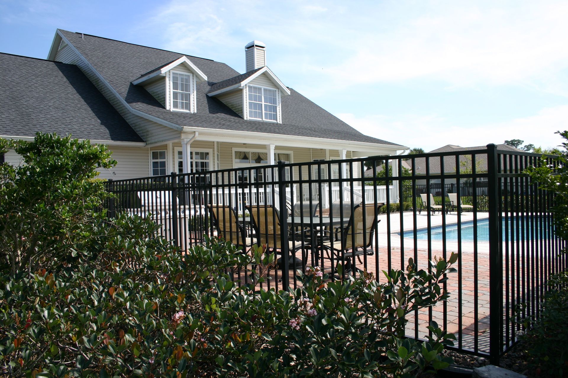 A cream-colored house with a dark roof and dormer windows overlooks a backyard pool area with a patio and black fence.