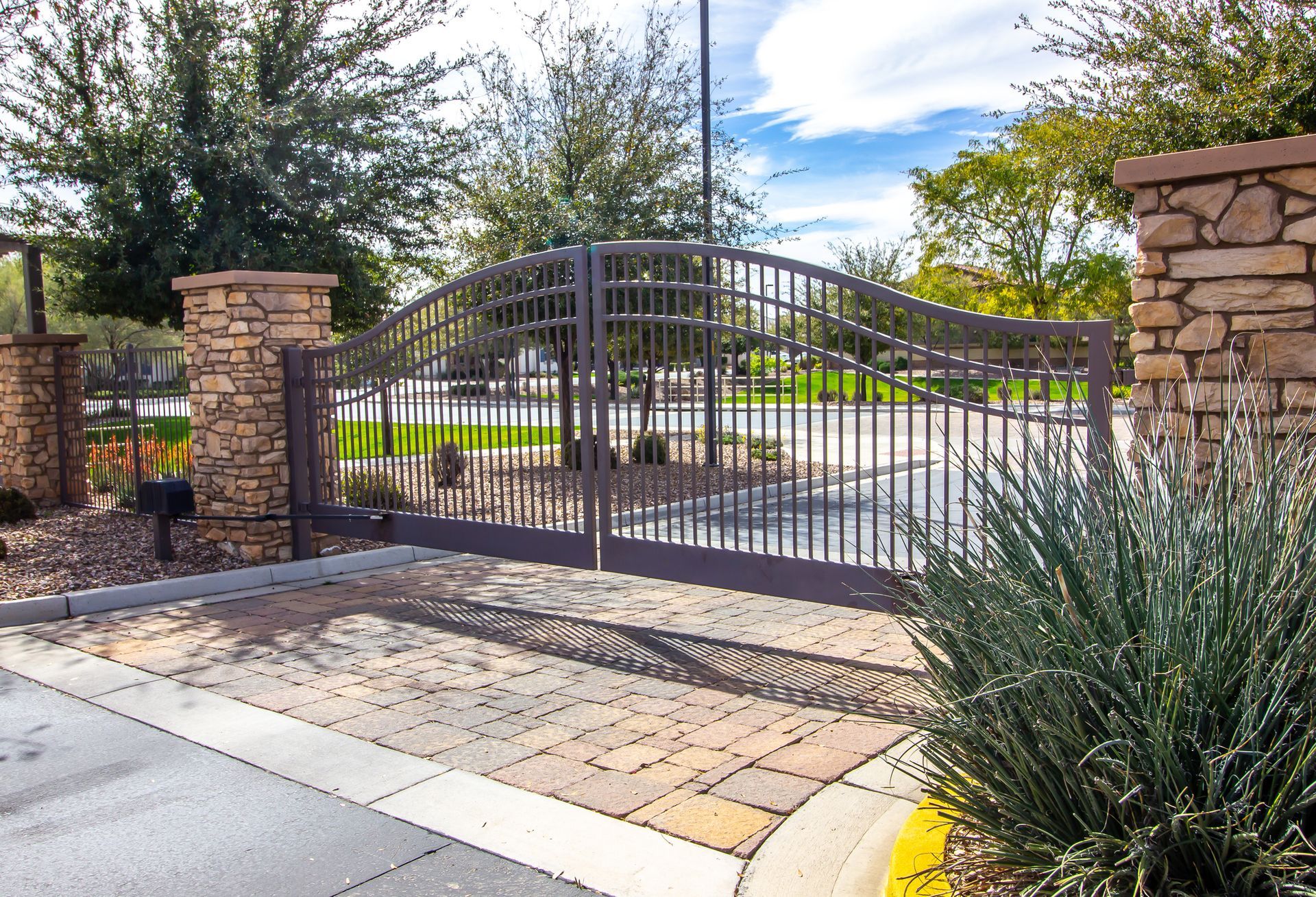 A closed dark metal gate with a curved top stands between two stone pillars at a paved entrance under a sunny sky.