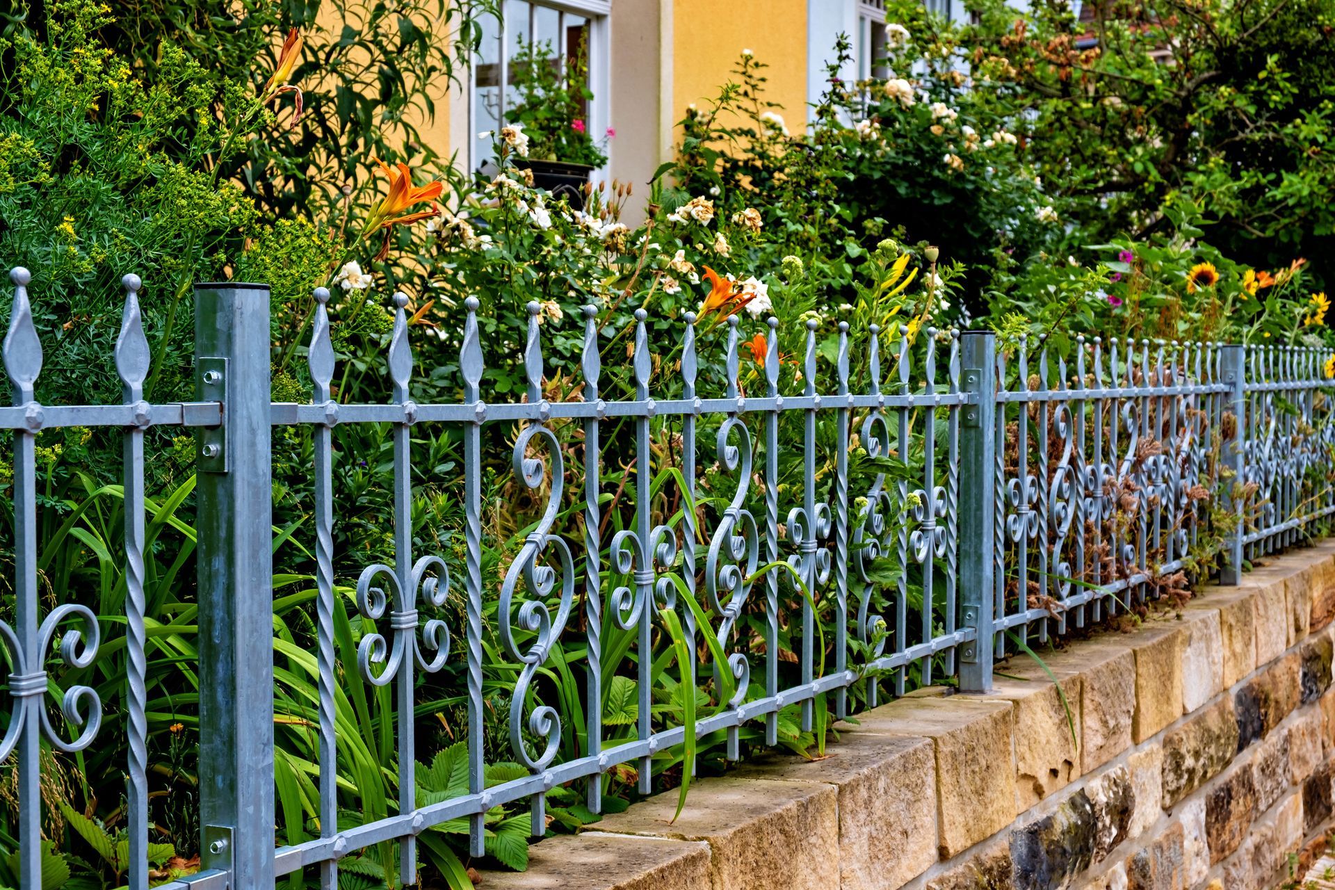 A metal fence with decorative, twisted uprights and spear finials, mounted on a stone wall in front of a garden.