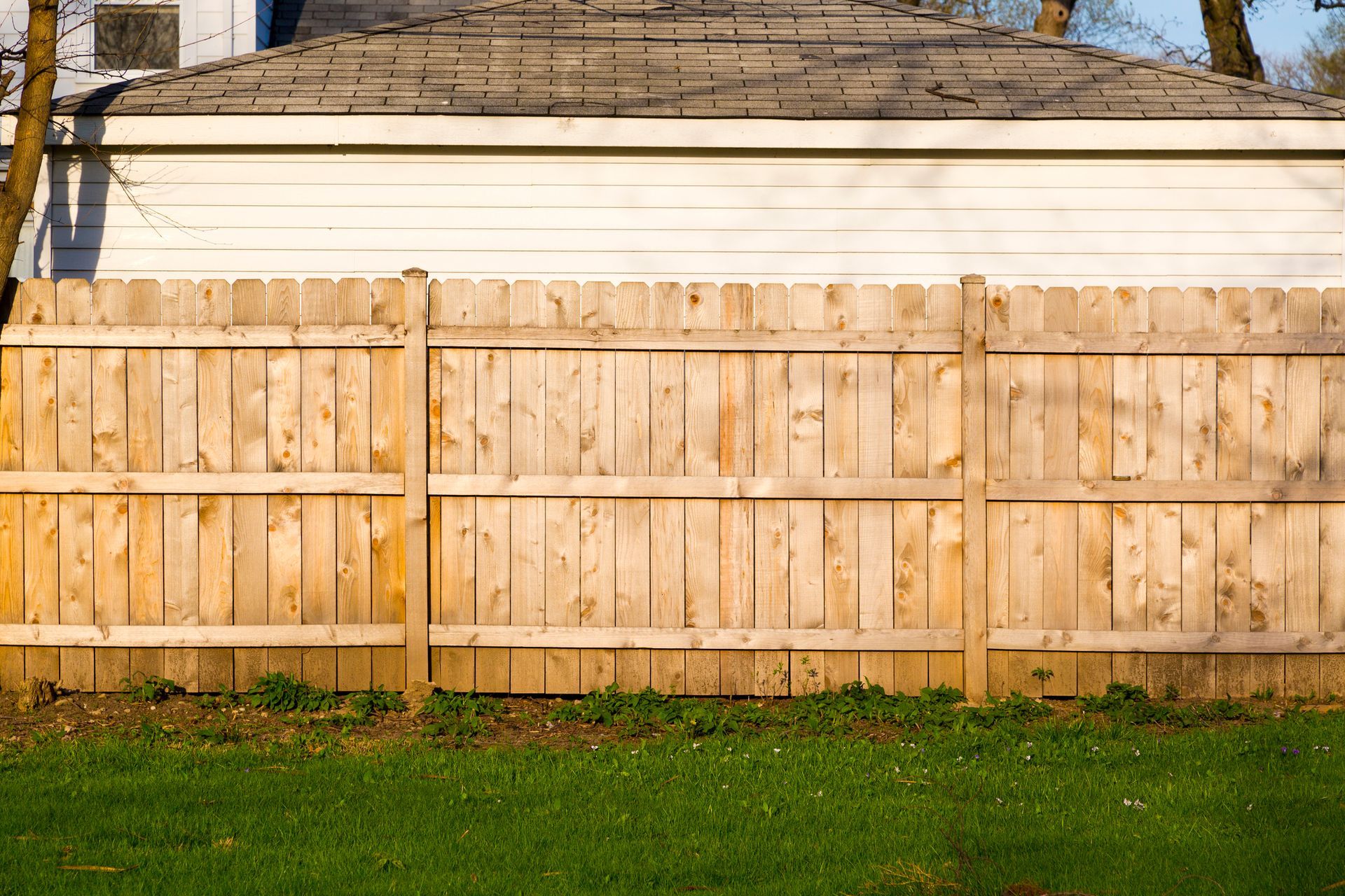A horizontal wood fence in front of a white building with a gray roof, set against a green lawn.