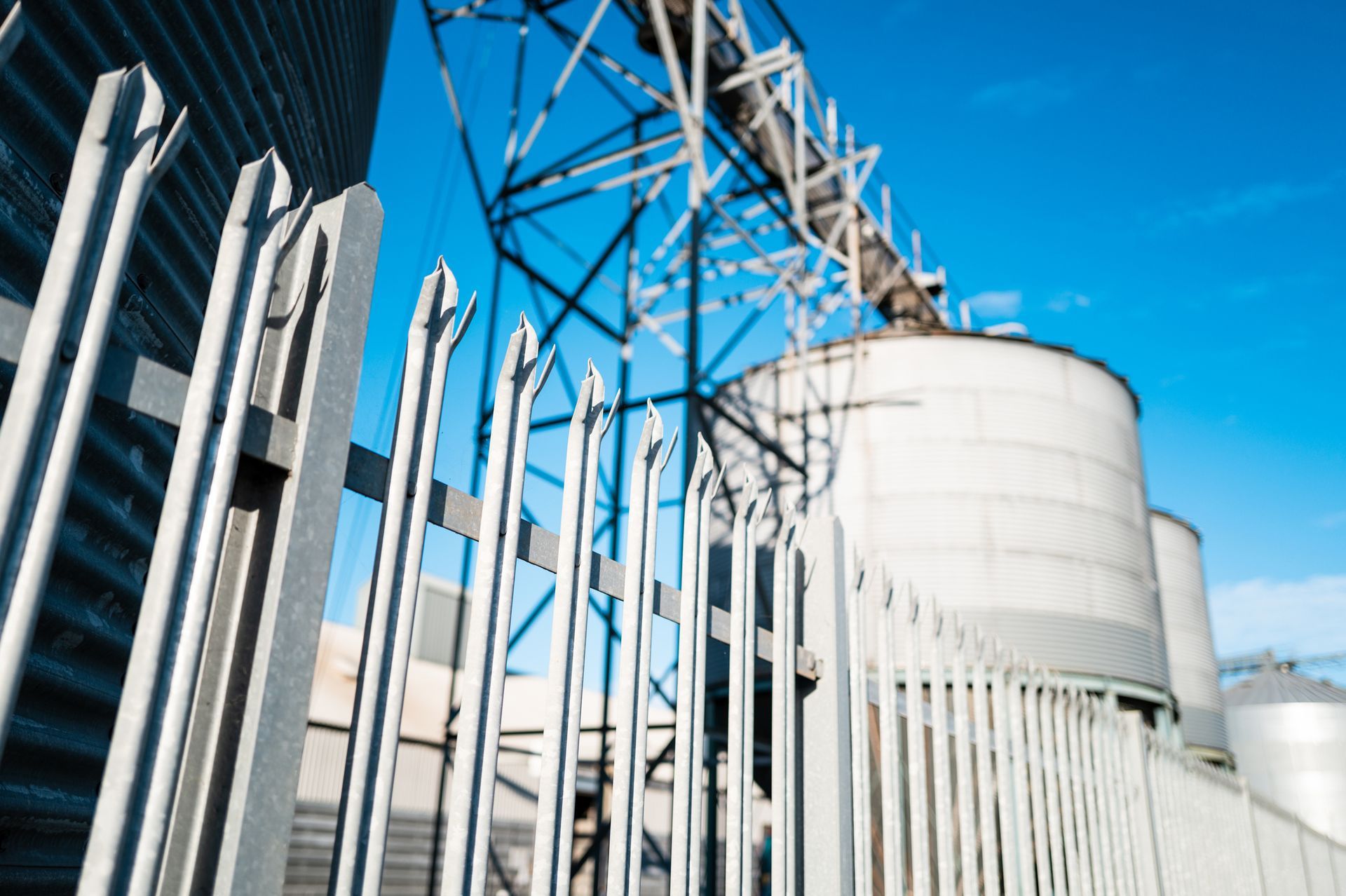 A metal fence in the foreground sits before large industrial silos and a tall steel structure against a clear blue sky.