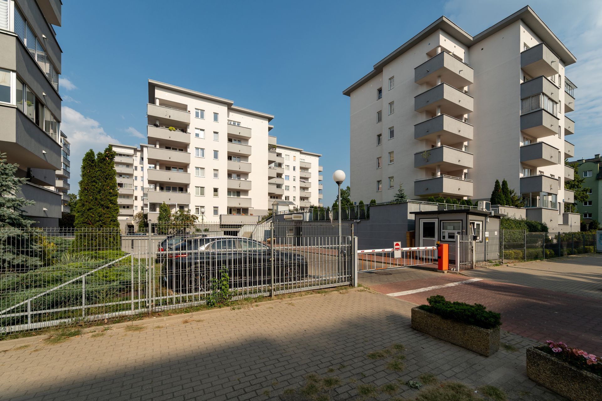 Modern apartment buildings with gray balconies behind a gated entrance with a security booth and metal fence.