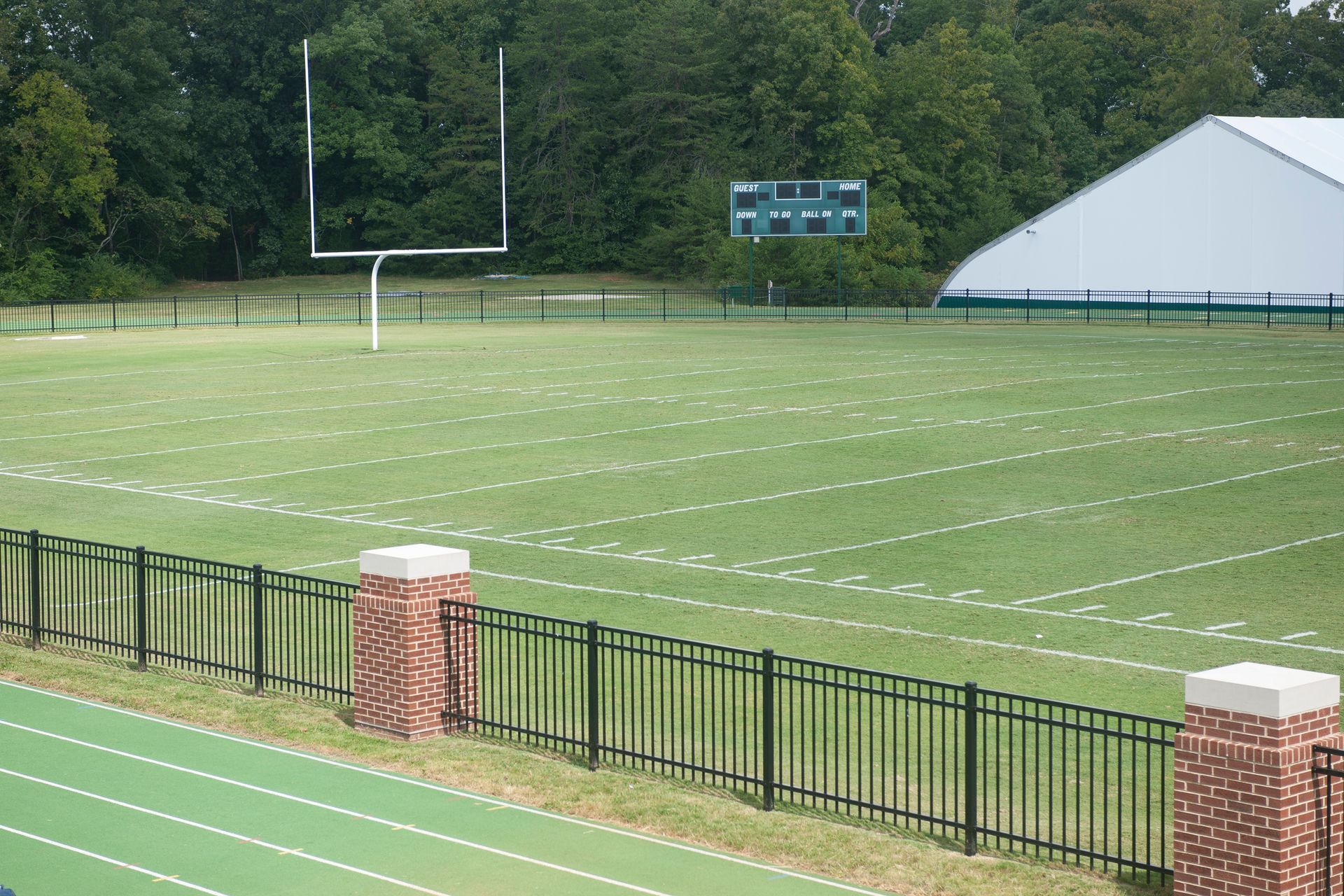 A football field with white yard lines, a goal post, and a scoreboard, viewed from a track behind a brick and metal fence.