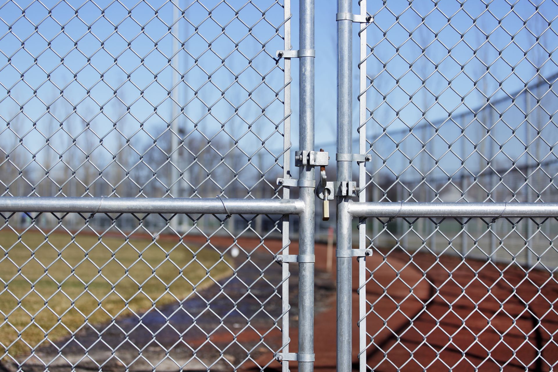 A chain-link fence gate closes off a field with a reddish track and green grass under a clear blue sky.