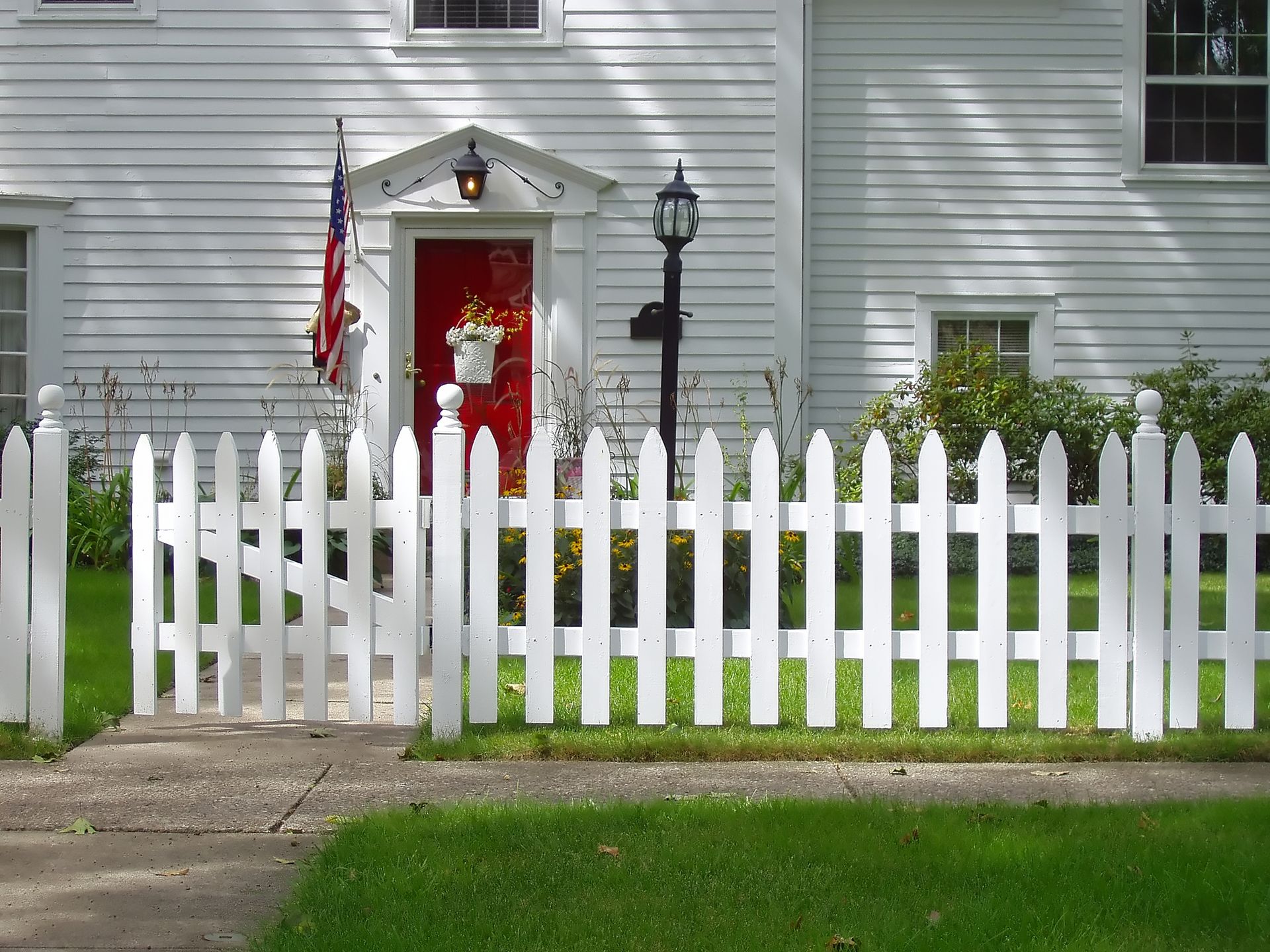 A white picket fence stands in front of a colonial-style house with white siding and a bright red front door.