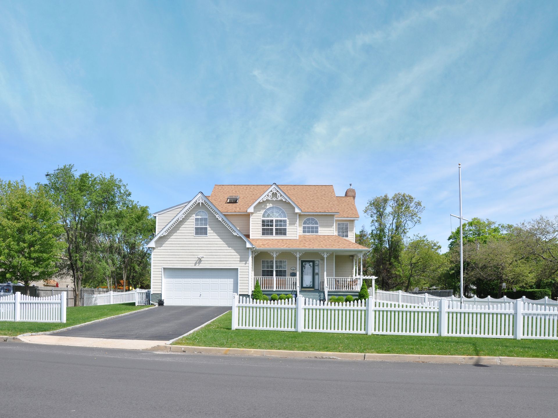A light-colored two-story house with a white garage and porch behind a white picket fence under a blue sky.